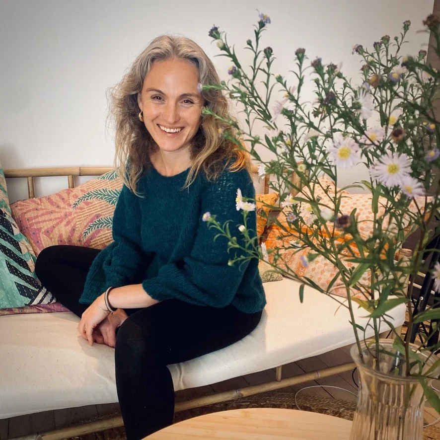 Kvinde sidder på en seng med farverige puder, smiler og har blomster i en vase foran sig.