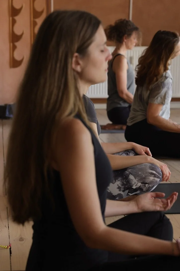 Fire kvinder sidder i lotusstilling med lukkede øjne i et yogarum.