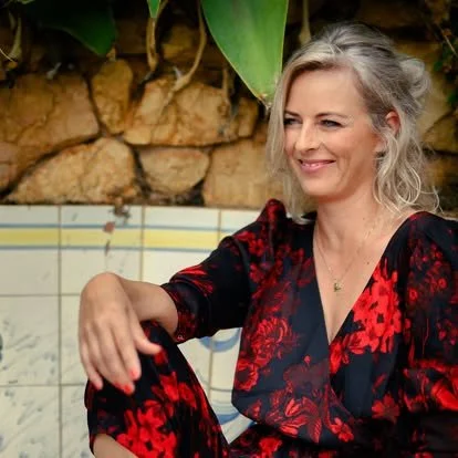 Kvinde med blond hår i en rød og sort blomsterdrenget kjole, smilende, sidder udenfor, baggrund med mursten og store grønne blade.