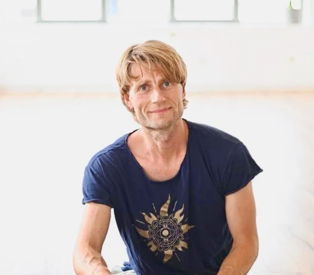 Mand sidder på gulvtæppet i et lyst rum med store vinduer bag ham, iført en blå t-shirt med et biologisk motiv.