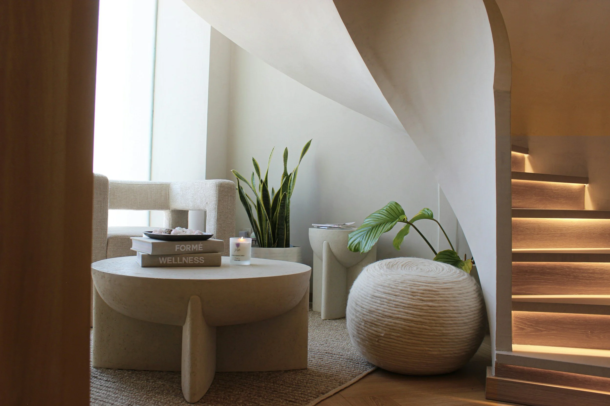 Modern living room with a beige armchair, green plants, a round stone coffee table, stacked books, a lit candle, and wooden stairs with built-in lighting.
