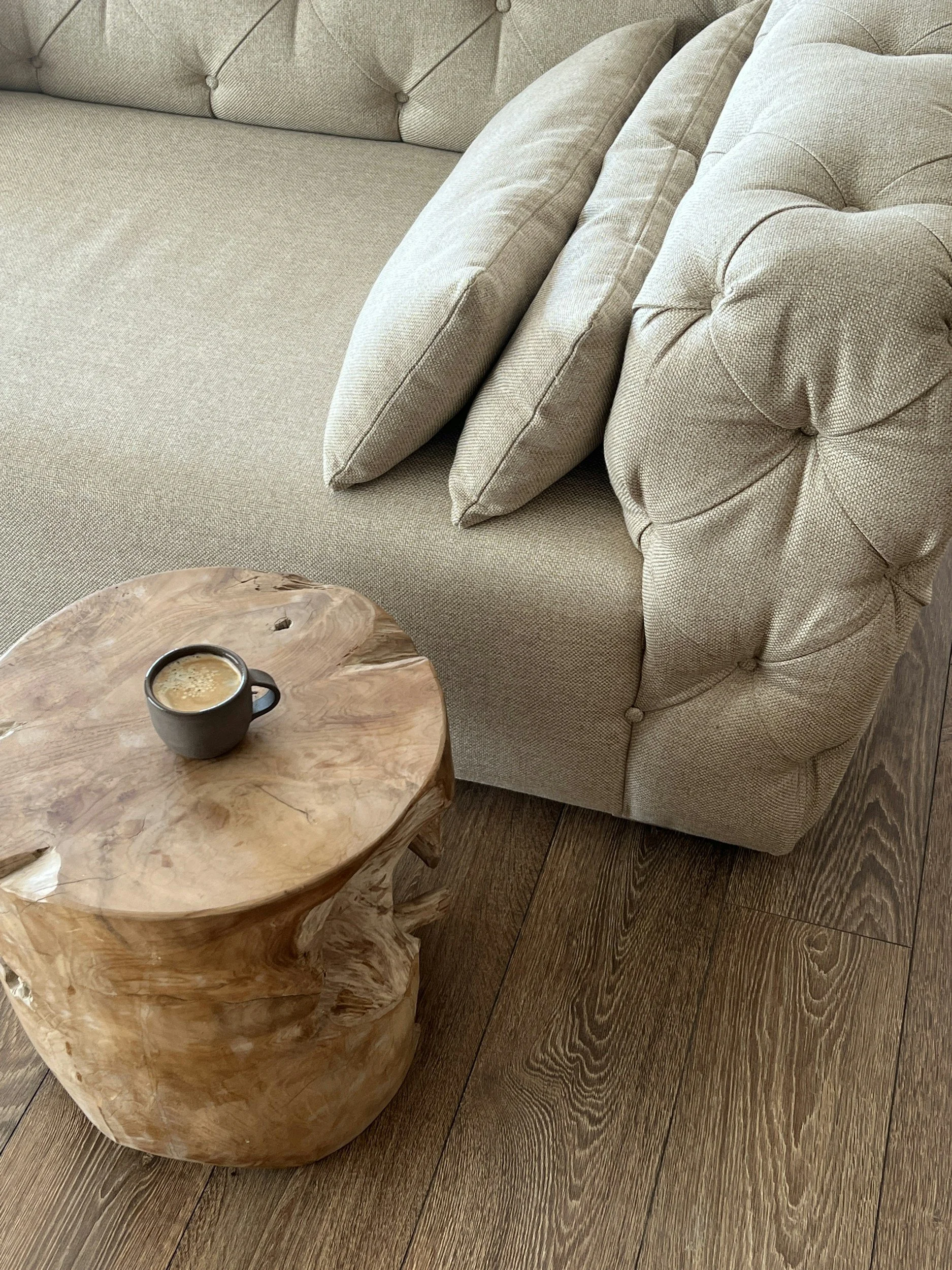 A beige sofa with rolled arms and several matching cushions, adjacent to a natural wood side table with a cup of coffee on it, set on a wooden floor.