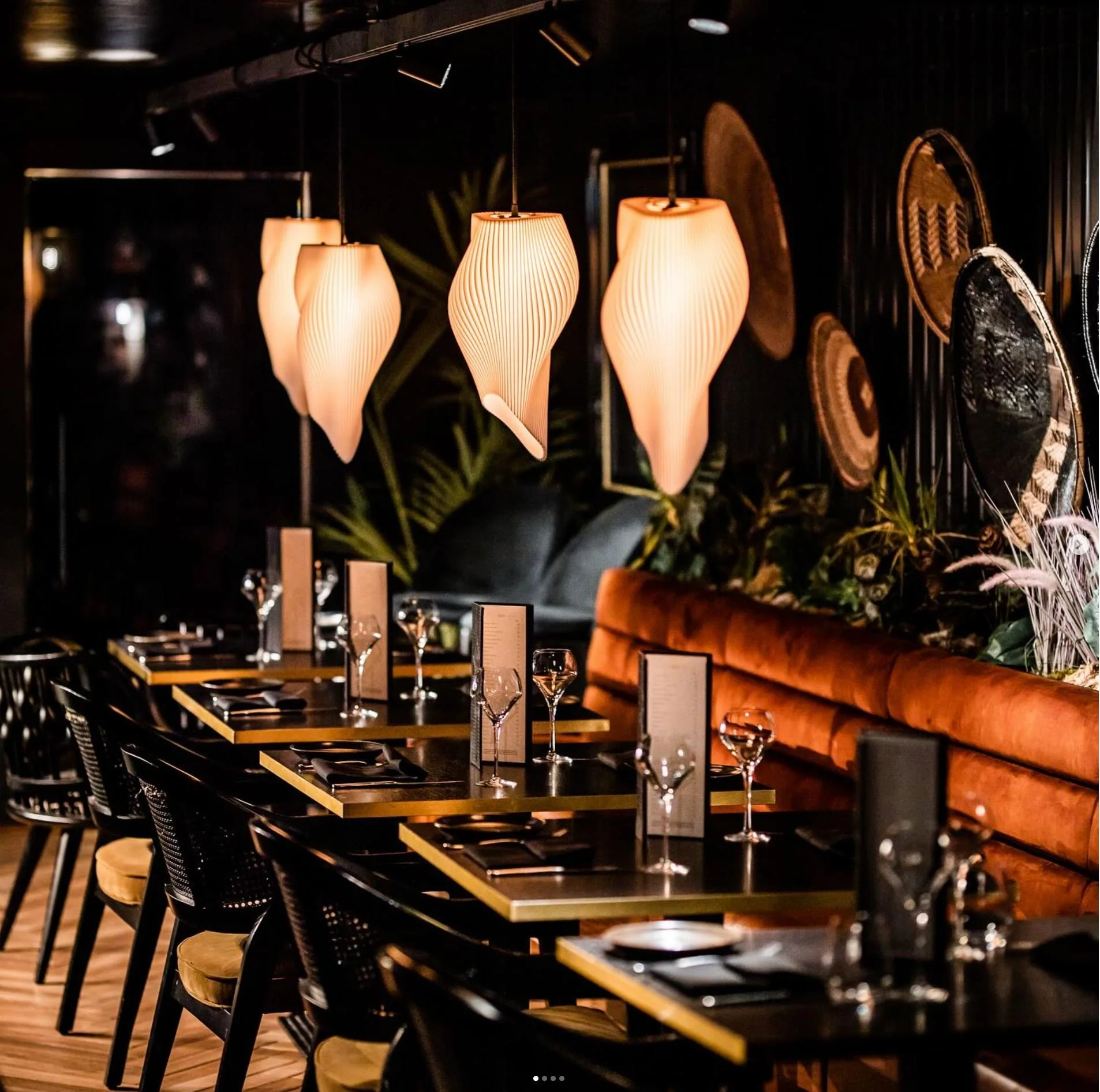 Elegant restaurant interior with a row of black tables set with wine glasses and menus, black chairs, and warm lighting from hanging pendant lamps. The decor includes a dark wall with woven art pieces and lush greenery.