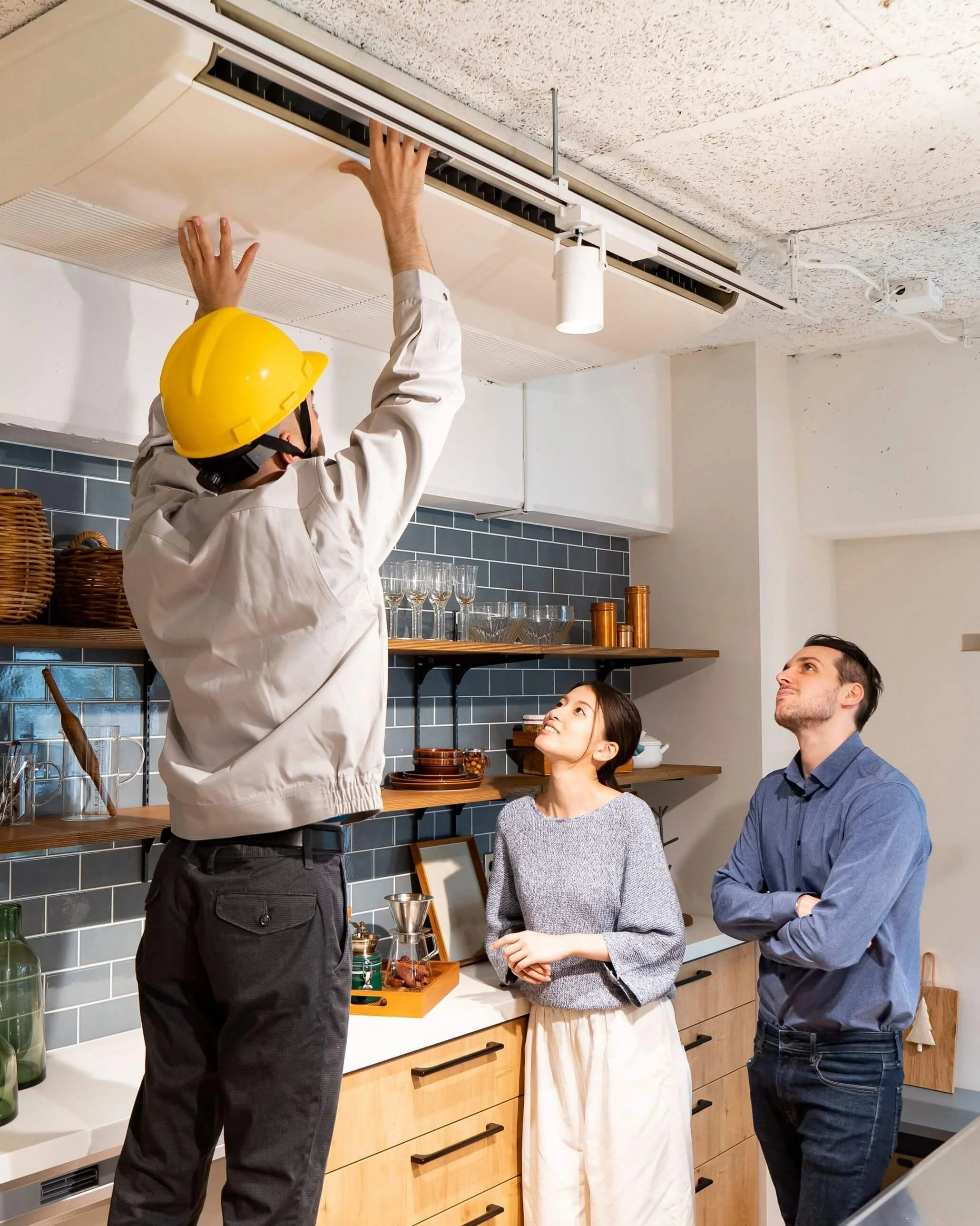 A man in a yellow safety helmet and white shirt is adjusting or repairing an air conditioning unit on the ceiling while three people watch and listen in a modern kitchen.
