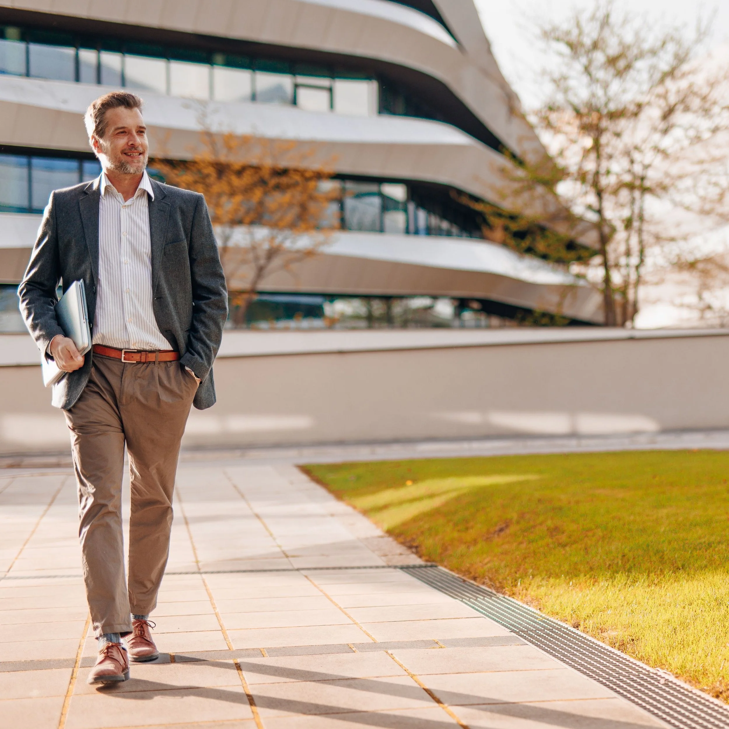 A man walking outdoors in front of modern apartment buildings. He has short hair, facial hair, and is dressed in a suit jacket, striped shirt, and brown pants, holding a laptop under his arm. Fall foliage is visible on the trees and ground.