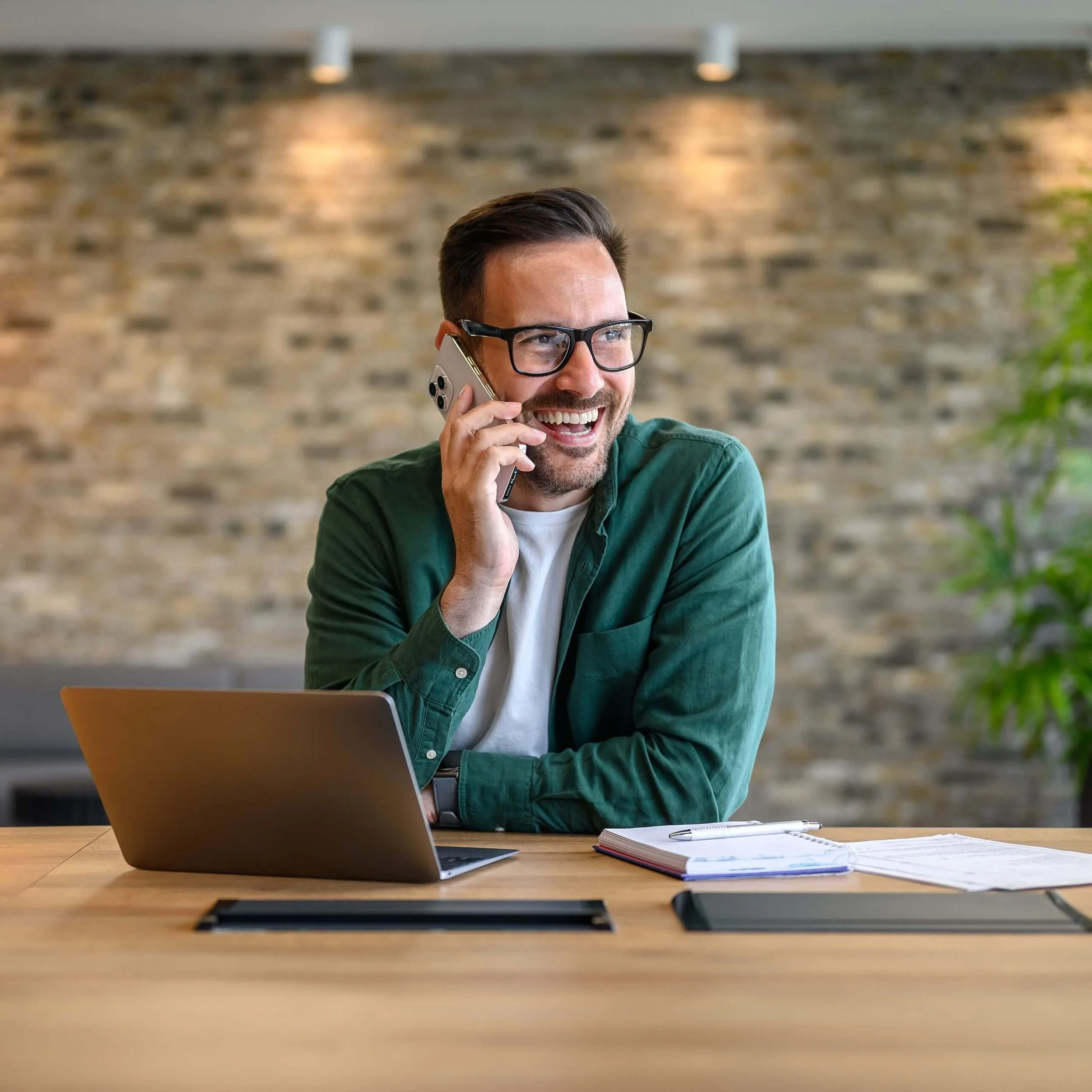 A man with glasses talking on a cell phone, sitting at a wooden desk with a laptop, notebook, and pen, in a modern office with a brick wall background.