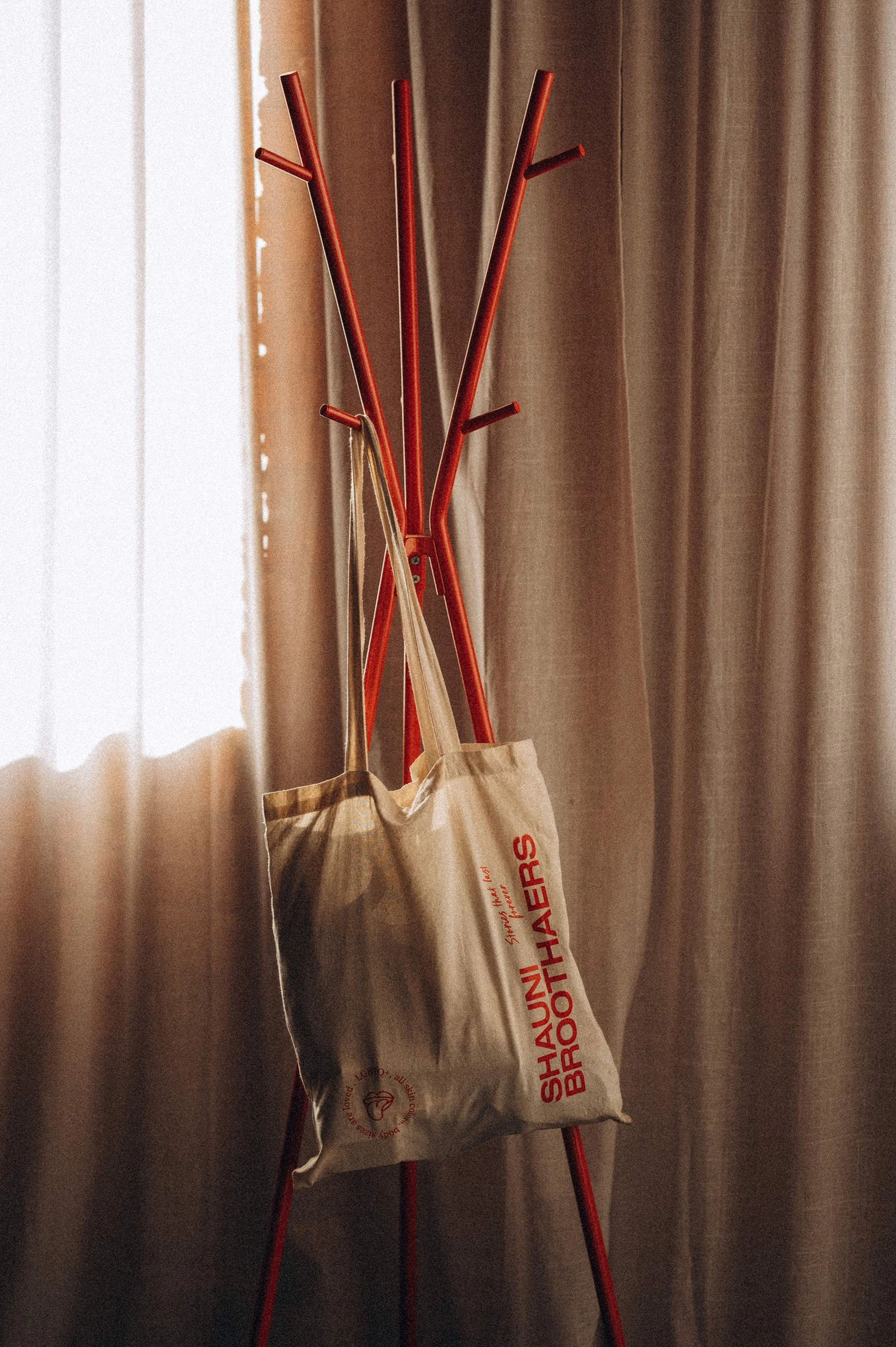 a tote bag with the shauni broothaers logo on it in red on a red coat rack with linnen curtains behind it