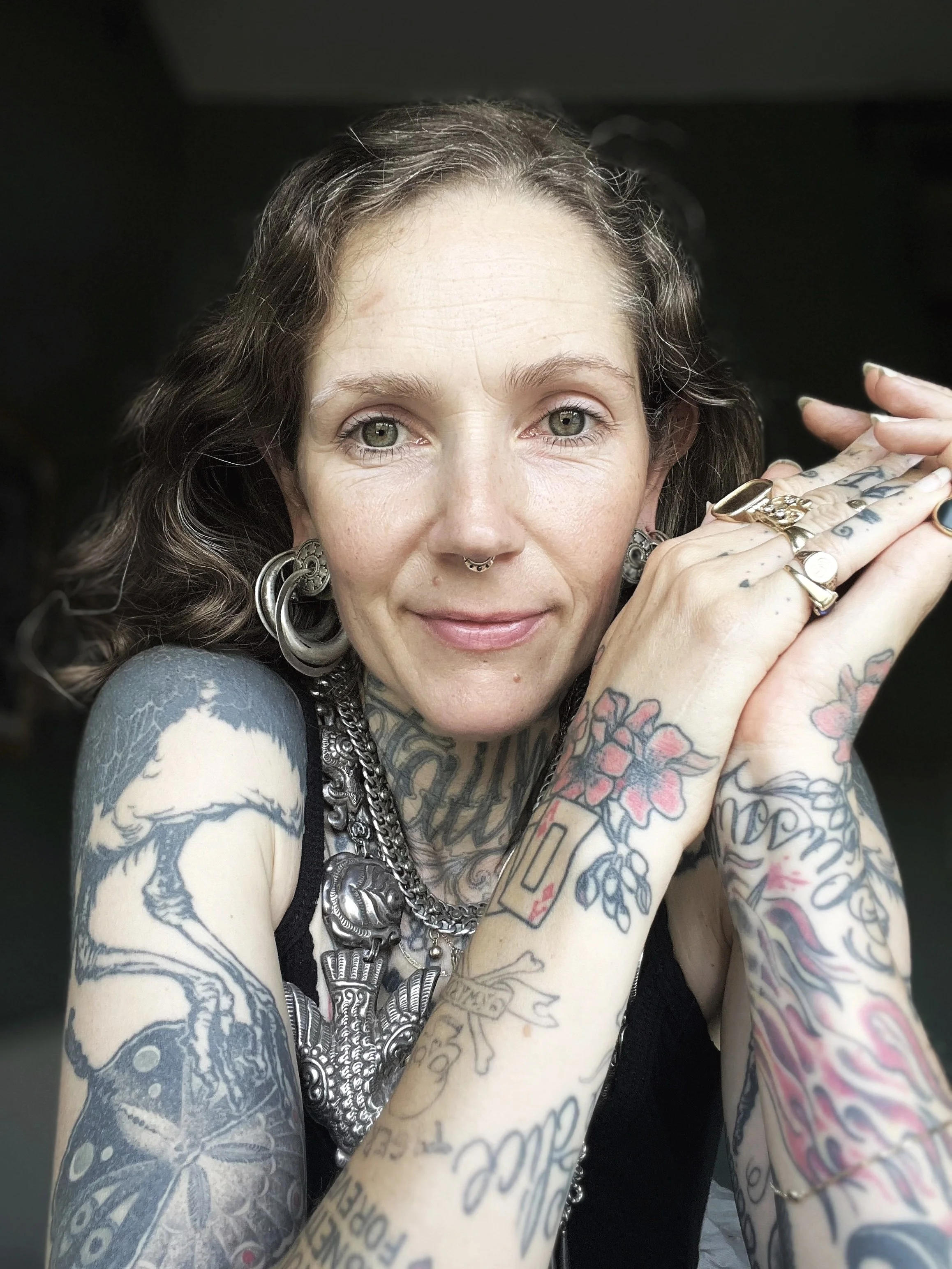 Close-up of Minka Sicklinger, a woman with tattoos, jewelry, and a septum piercing, resting her chin on her hands.