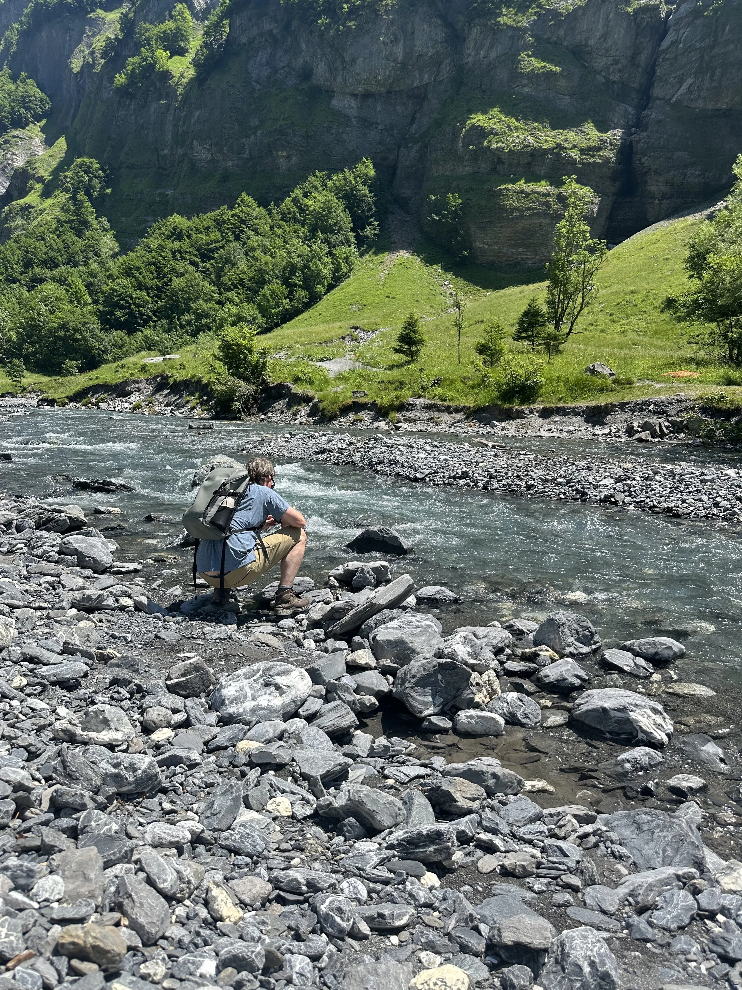 Person sitting on rocks by a river in a mountainous area, with green trees and cliffs in the background.