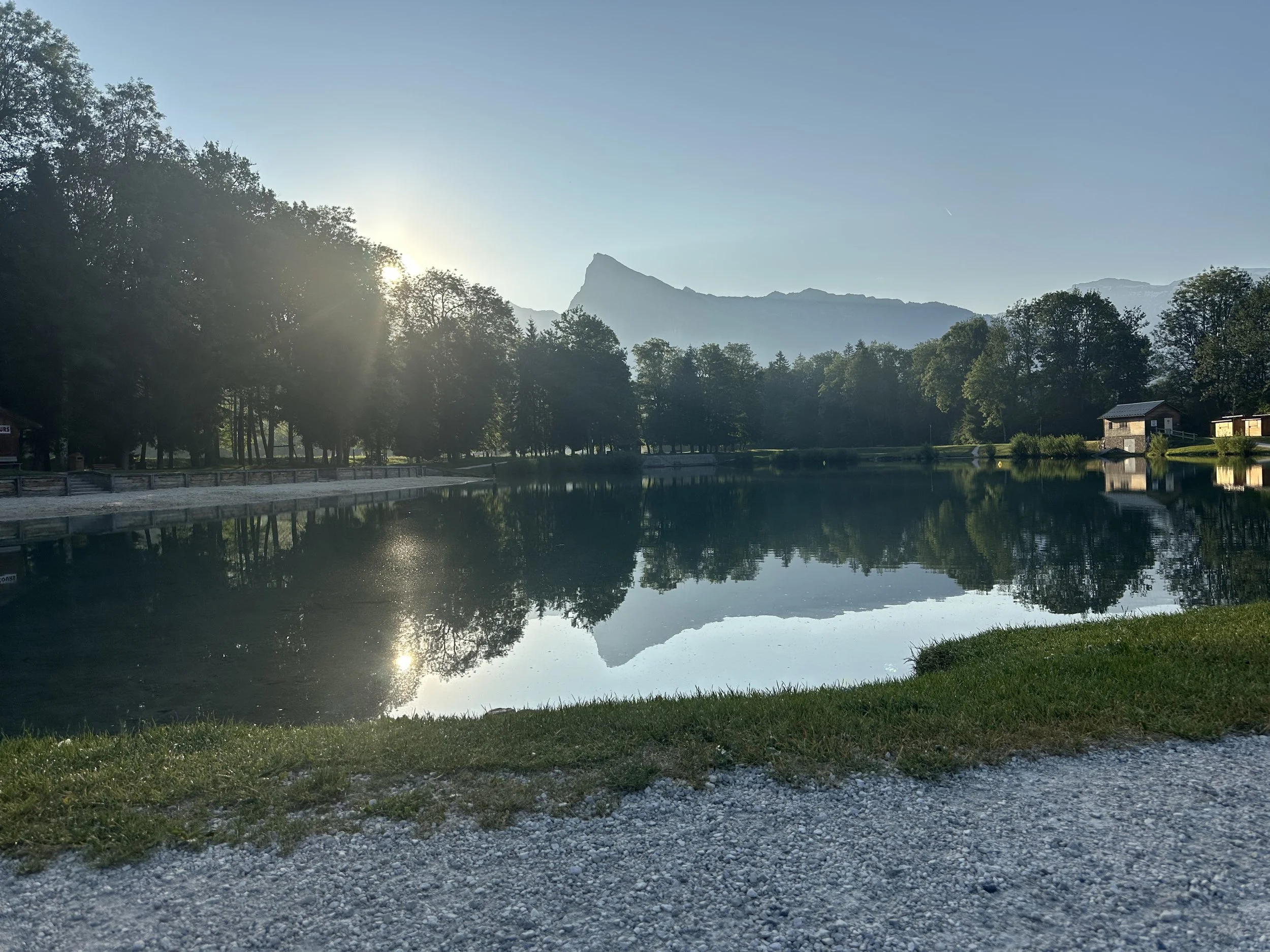 A peaceful lake scene at sunrise with mountain in the background, trees along the shore, and calm reflective water.