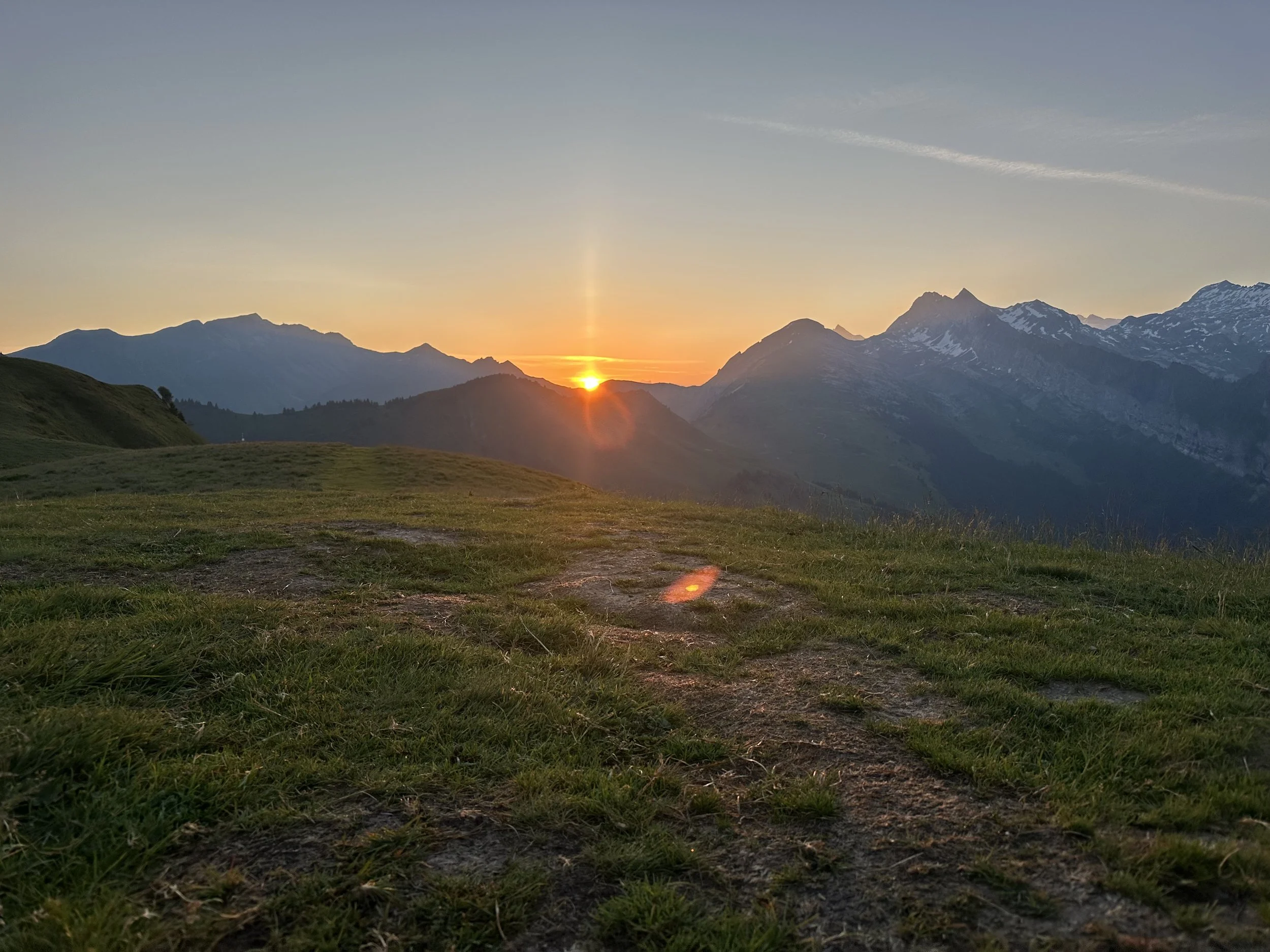 Sunset over green grassy hills and mountain range with partly cloudy sky.