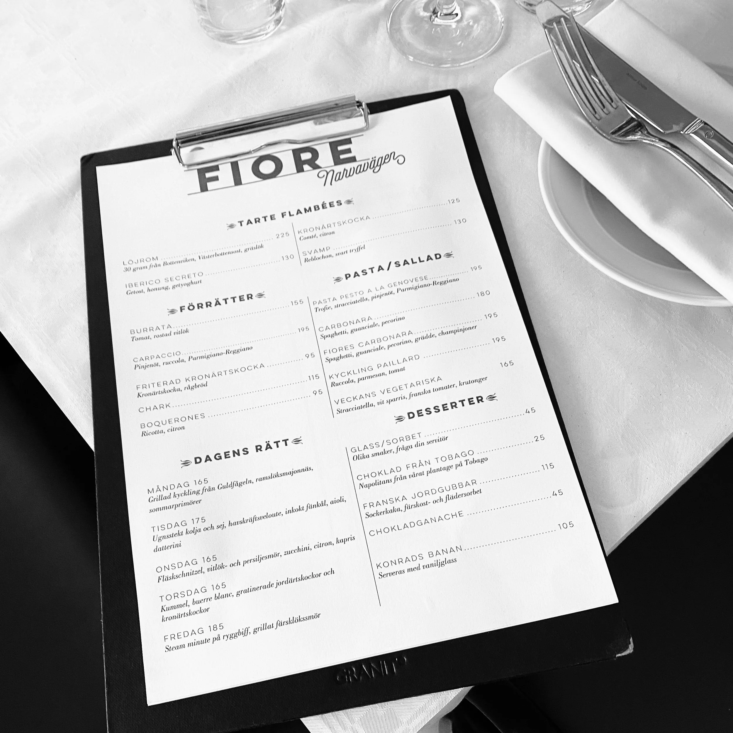 A restaurant menu on a clipboard with a white tablecloth, set with a dinner knife, fork, a napkin on a plate, and wine glasses in the background.