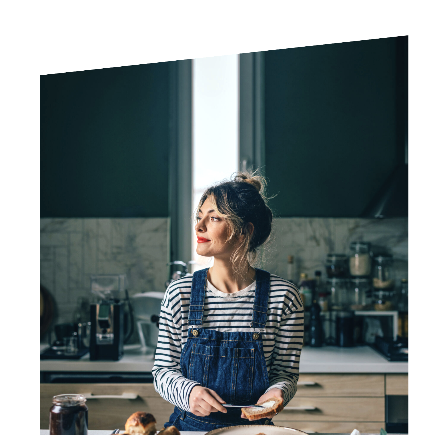Woman with blonde hair in a bun, wearing a striped shirt and denim apron, standing in a modern kitchen by a window.