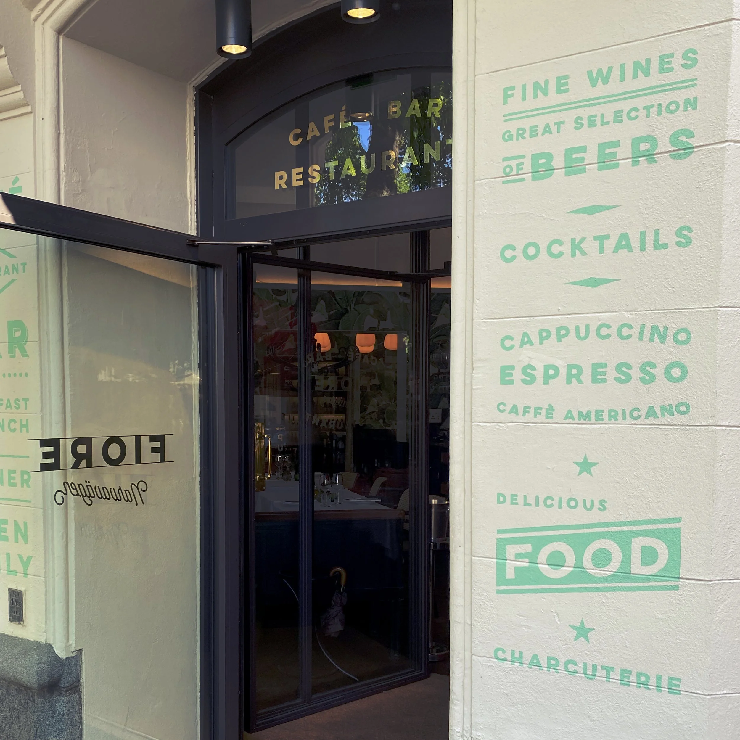 Exterior view of a restaurant with a glass door and wall painted with green text listing menu items like wines, beers, cocktails, cappuccino, espresso, and food, with a black awning above the door.