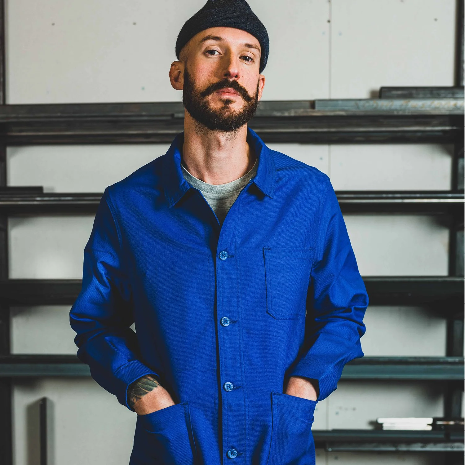 A man with a beard and mustache wearing a blue work jacket and a black beanie, standing with his hands in his pockets in front of a shelf with black metal bars.