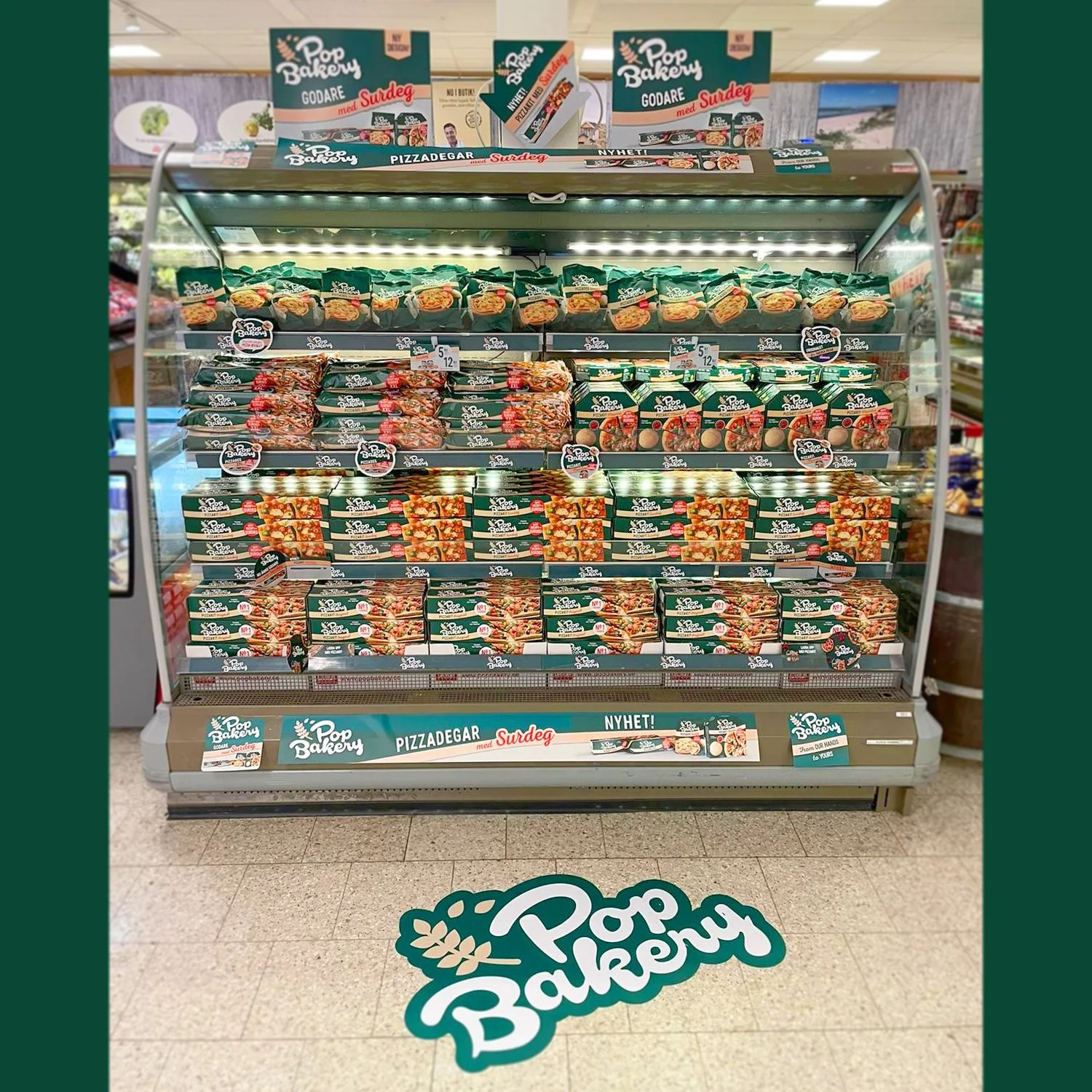 A refrigerated display in a store stocked with Pop Bakery frozen pizza products. The products are in green and white packaging, with images of pizza on the boxes. Signage on top and at the bottom promotes the brand and new product line, with the bran