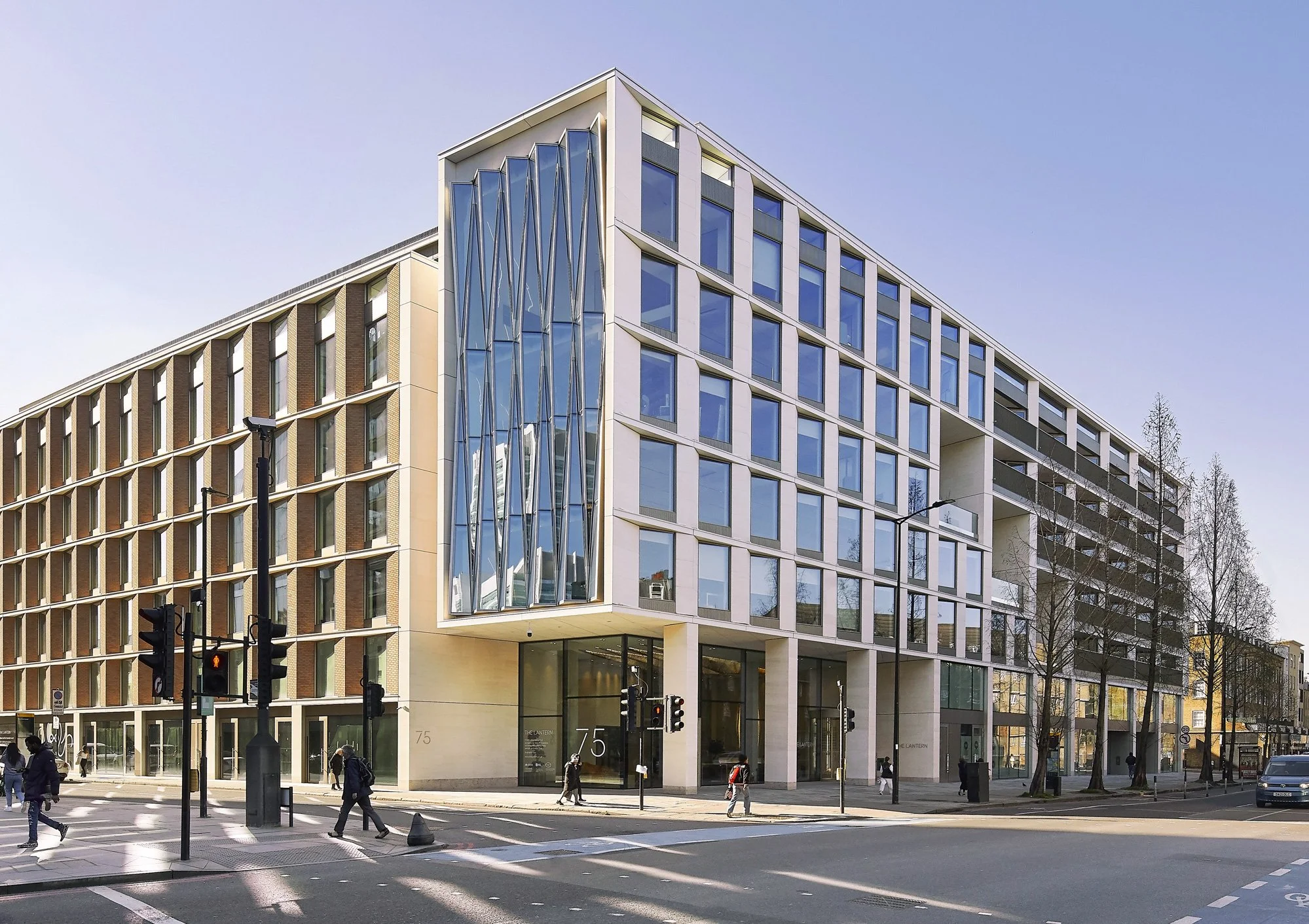 Modern multi-story building at street corner with glass and concrete facade, several pedestrians crossing the street, traffic lights, and leafless trees along the sidewalk.