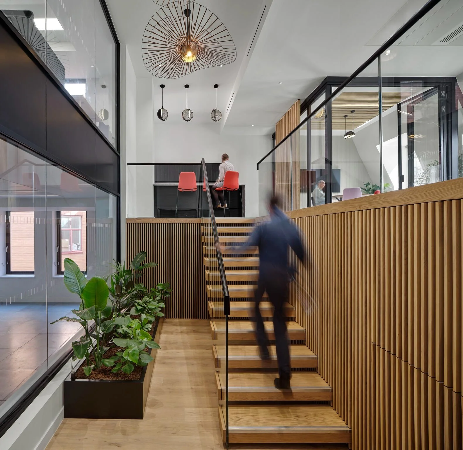 Modern interior with wooden stairs, glass railings, and plants; a person ascending stairs and two people seated upstairs in a contemporary office or co-working space.