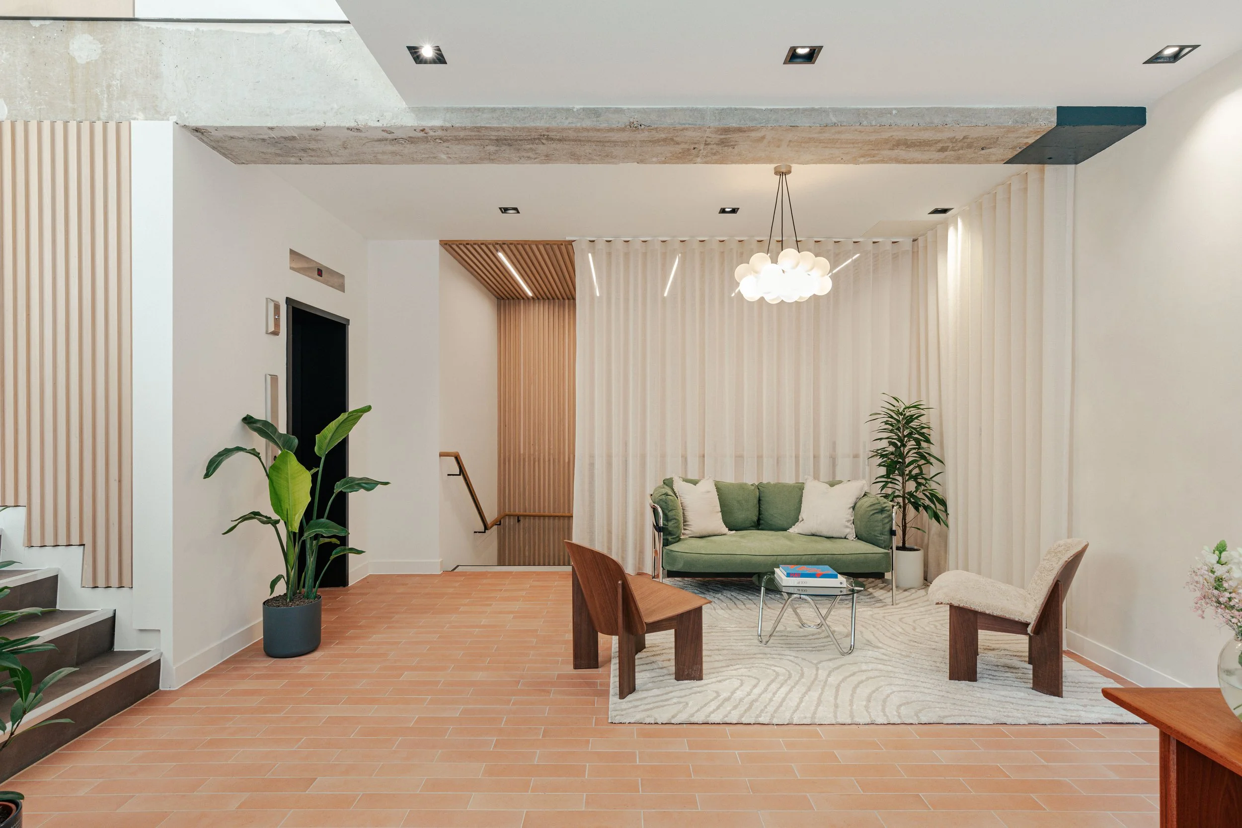 Modern lobby with a green sofa, decorative pillows, wooden and fabric chairs, potted plants, a rug, a ceiling chandelier, and curtains dividing the space.