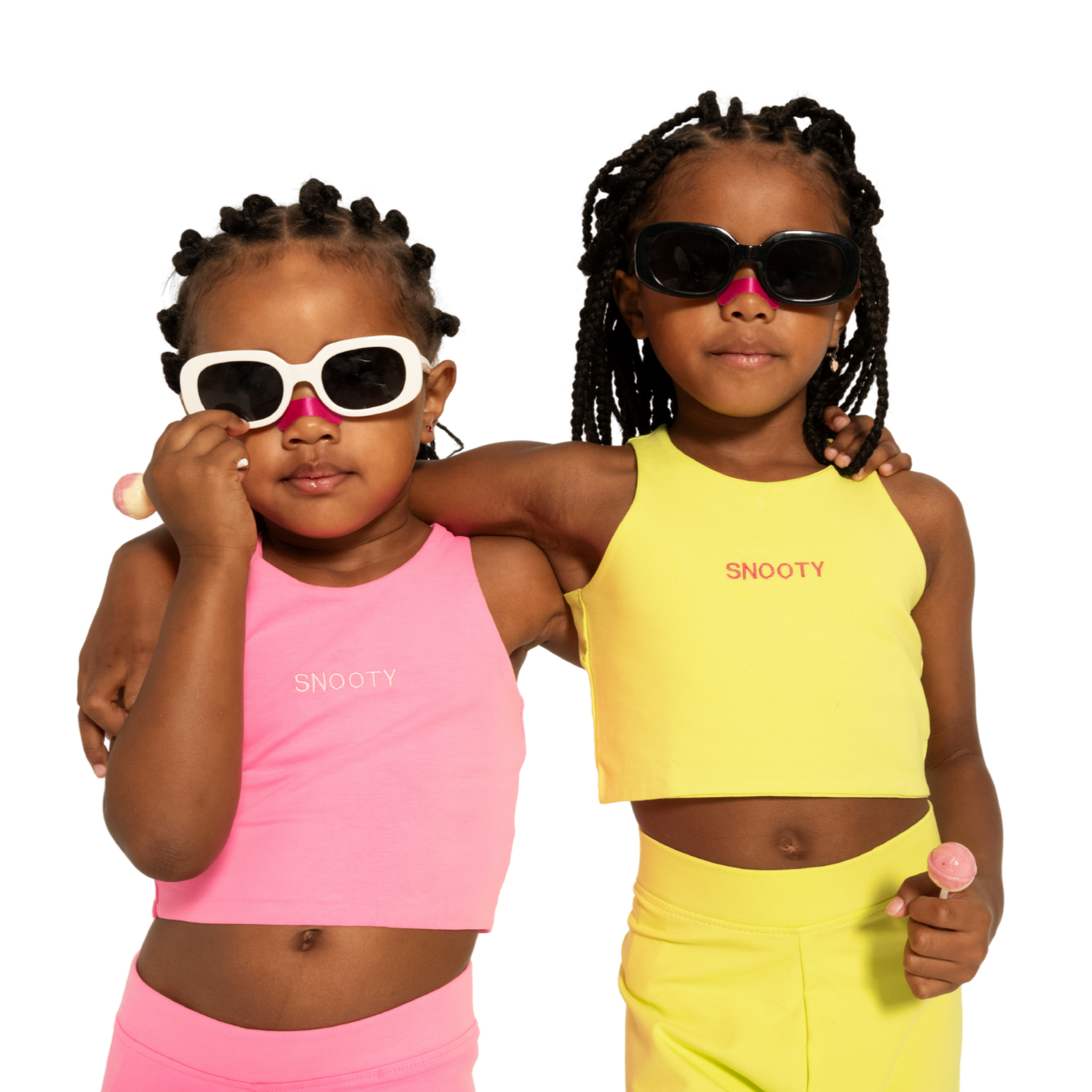 Zwei junge Mädchen mit Sonnenbrillen, eines in Pink, das andere in Gelb, stehen nebeneinander, umarmen sich, tragen Sportkleidung mit dem Label 'SNOOTY' und halten ein Lutscher.