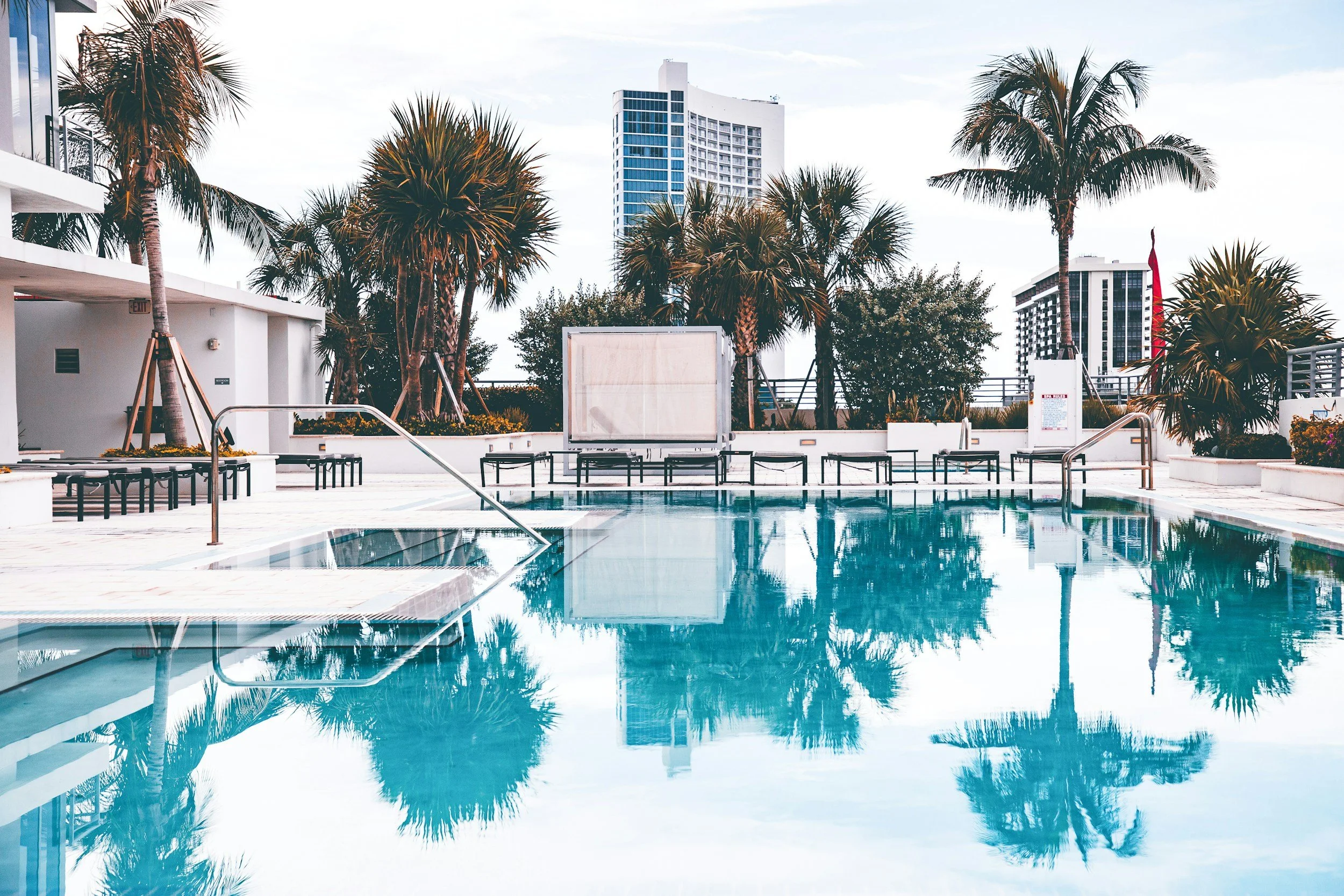 Empty outdoor swimming pool with palm trees and high-rise buildings in the background.