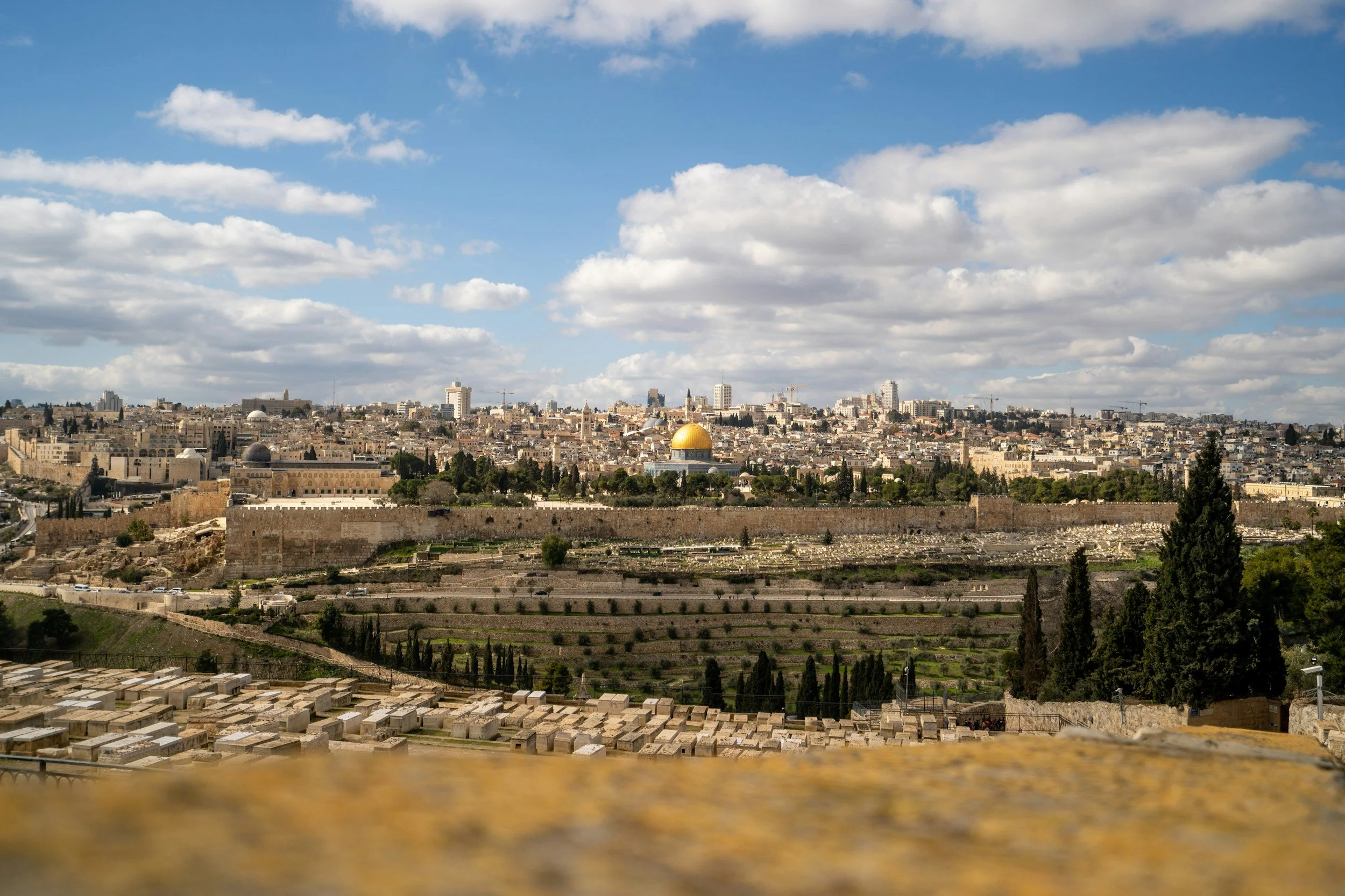 A cityscape of Jerusalem with the golden Dome of the Rock in the center, surrounded by ancient walls and modern buildings under a partly cloudy sky.