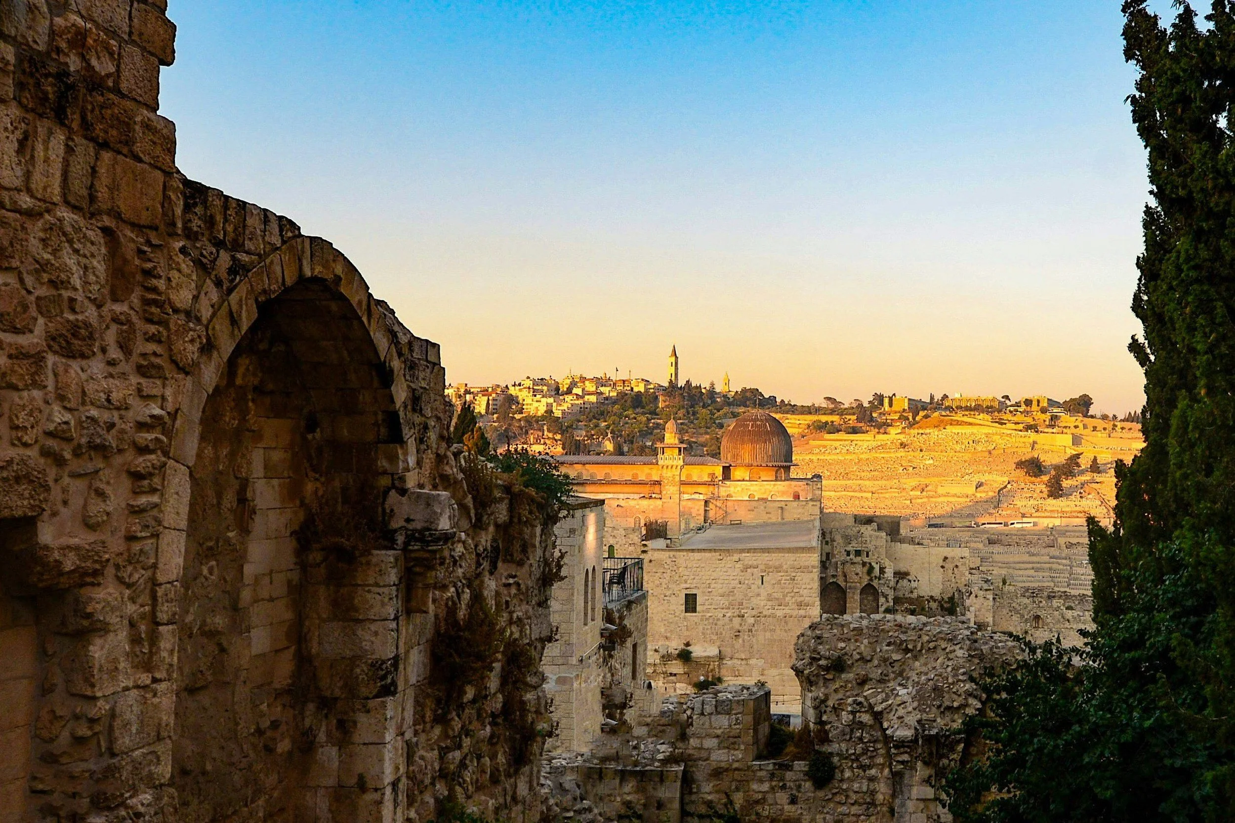 Sunset view of an ancient stone city with a domed building, a tower, and terraced hills in the background.