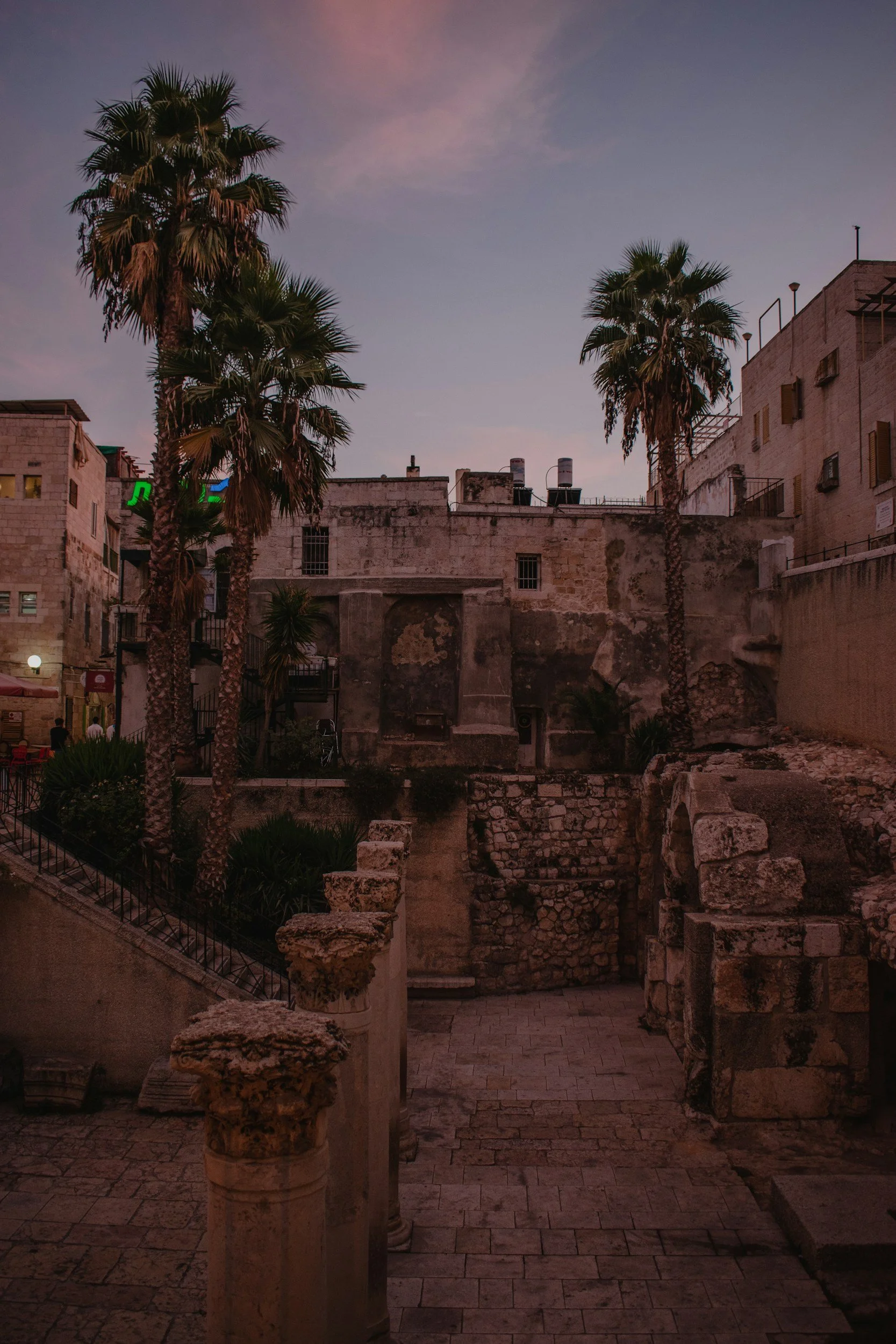 Old stone ruins and palm trees at sunset or dusk in an urban setting with buildings in the background.