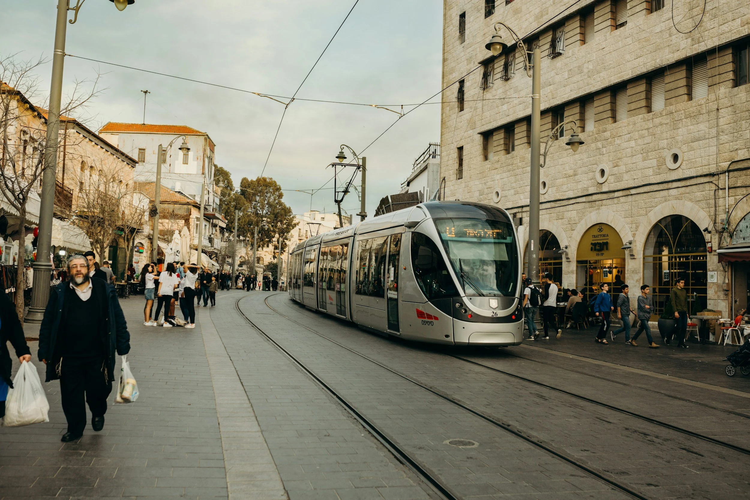 Modern tram on city street with pedestrians, storefronts, and historic buildings in the background.