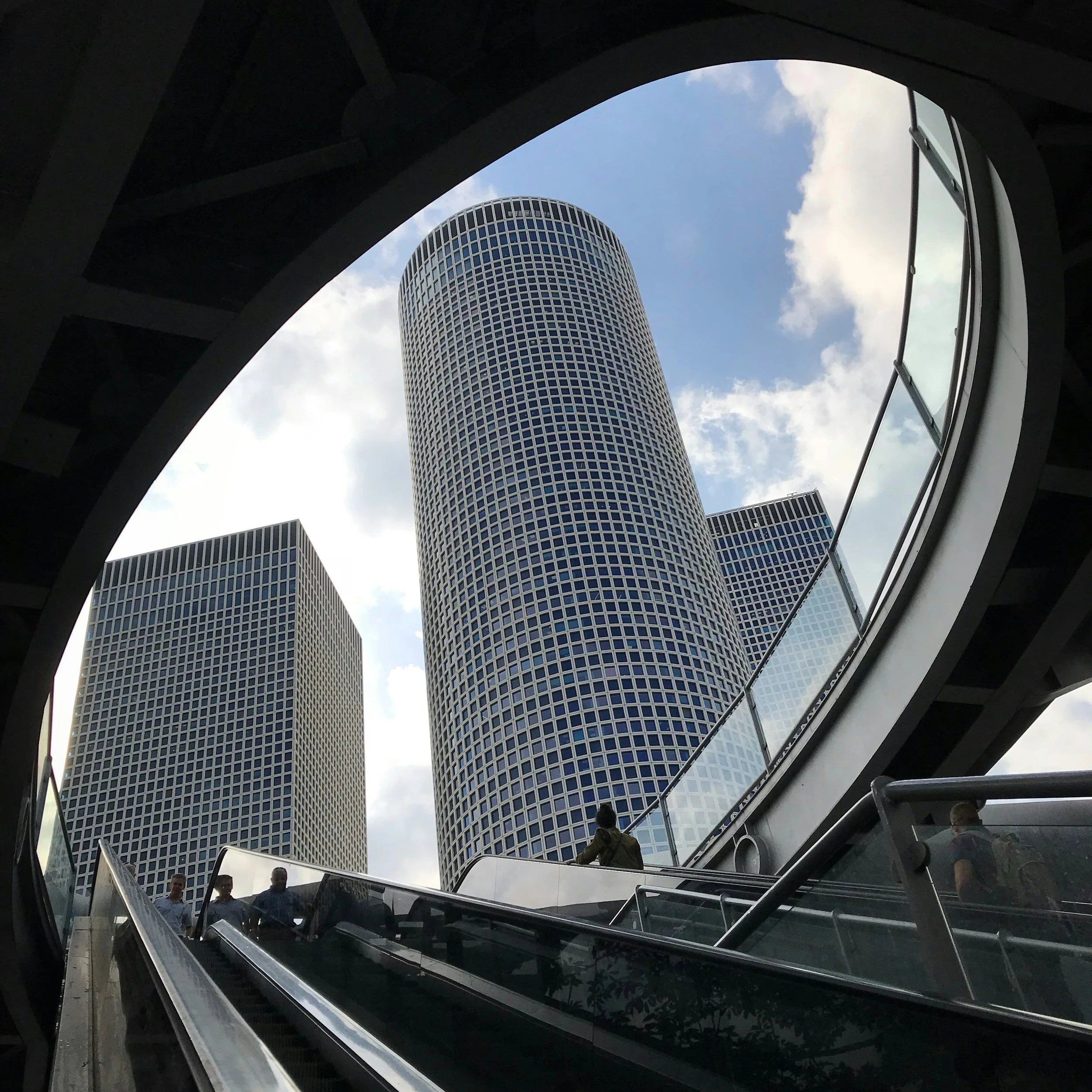 View of tall modern office buildings seen from an underground walkway with escalators, under a cloudy sky.