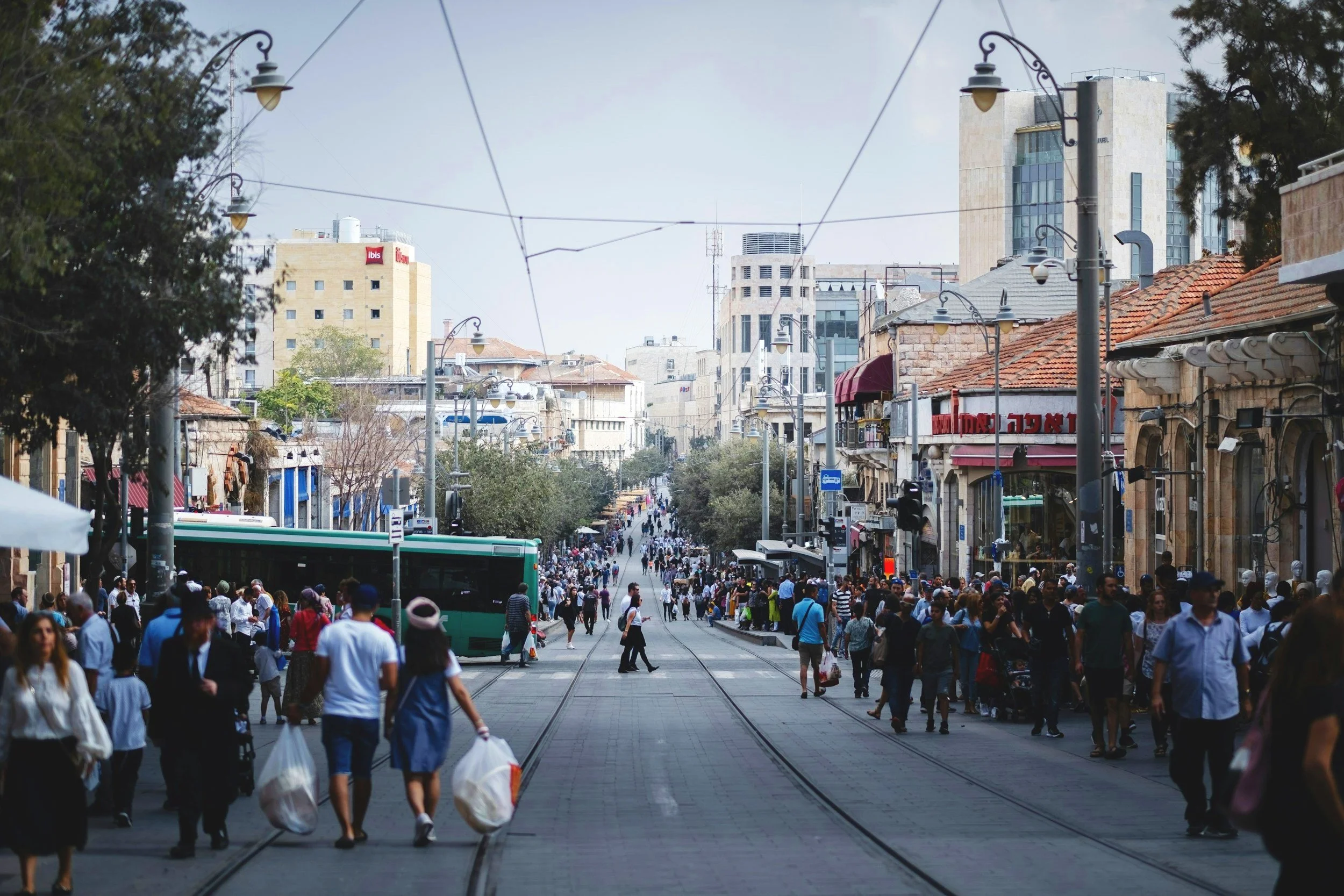 A busy city street with tram tracks, numerous pedestrians, shops on either side, and tall buildings in the background.