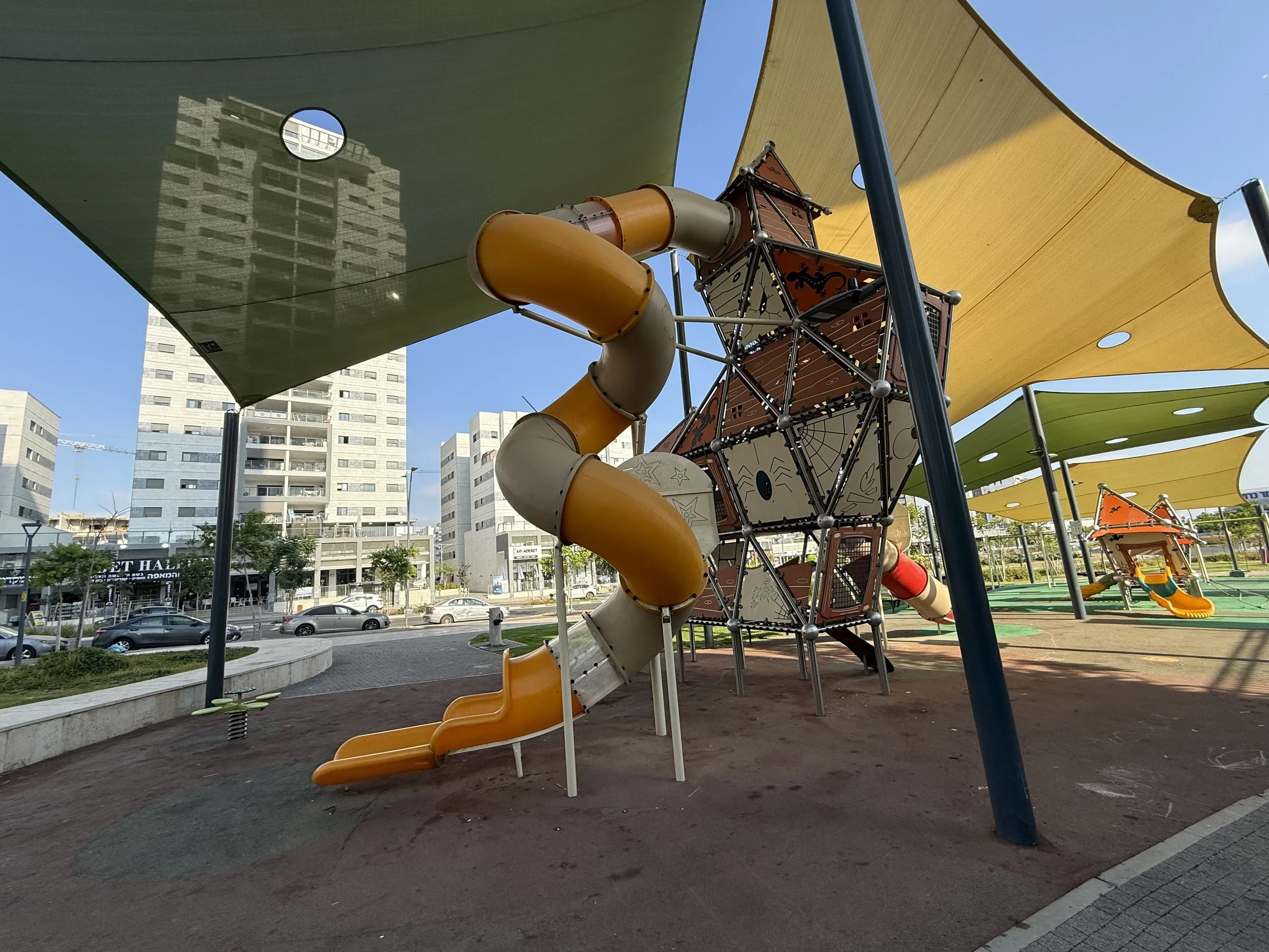 Children's playground with slides, climbing structures, and shade sail coverings in an urban park setting with buildings and parked cars in the background.