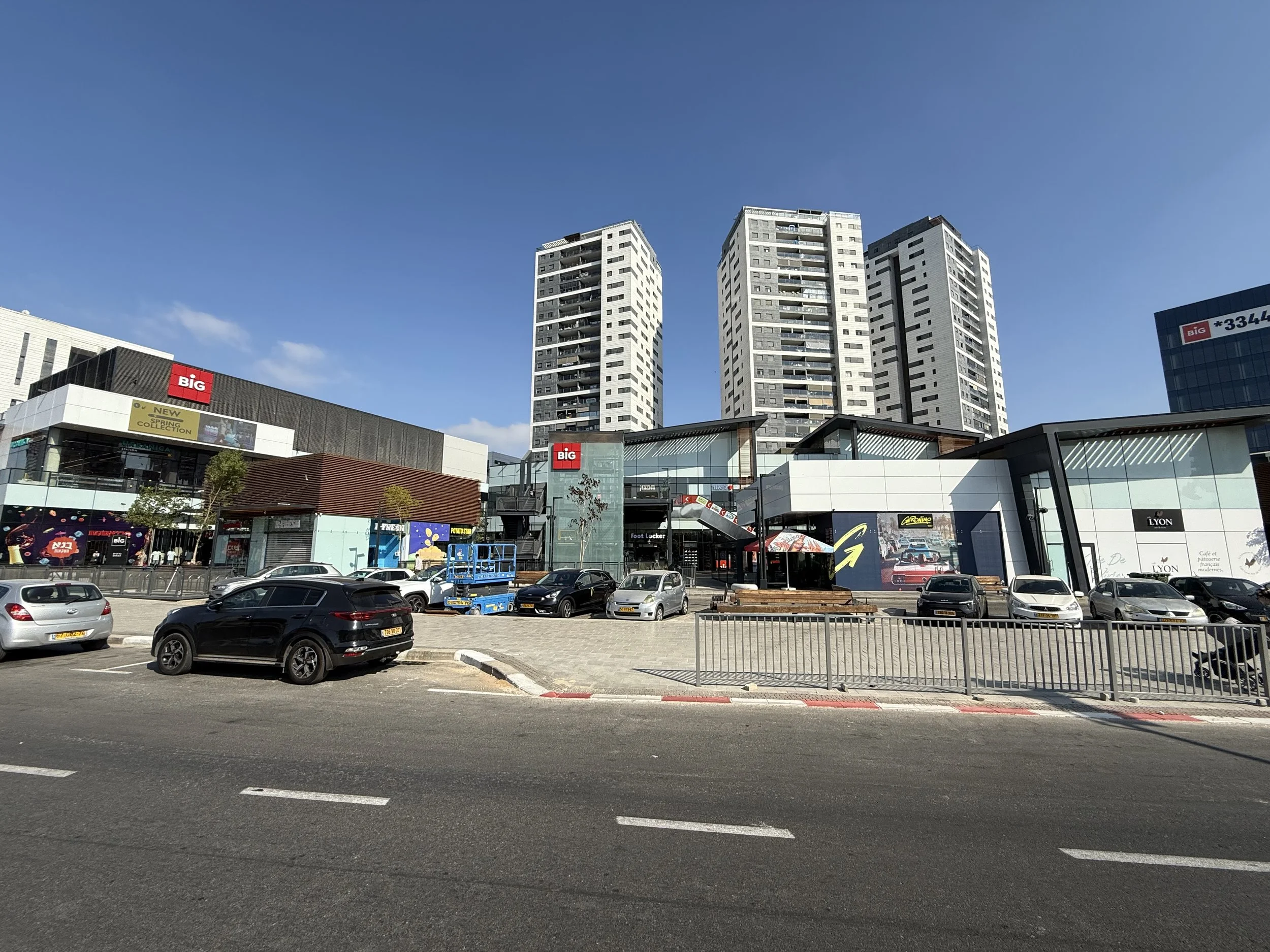 Shopping mall with stores, parking lot, and high-rise buildings in the background under a clear blue sky.