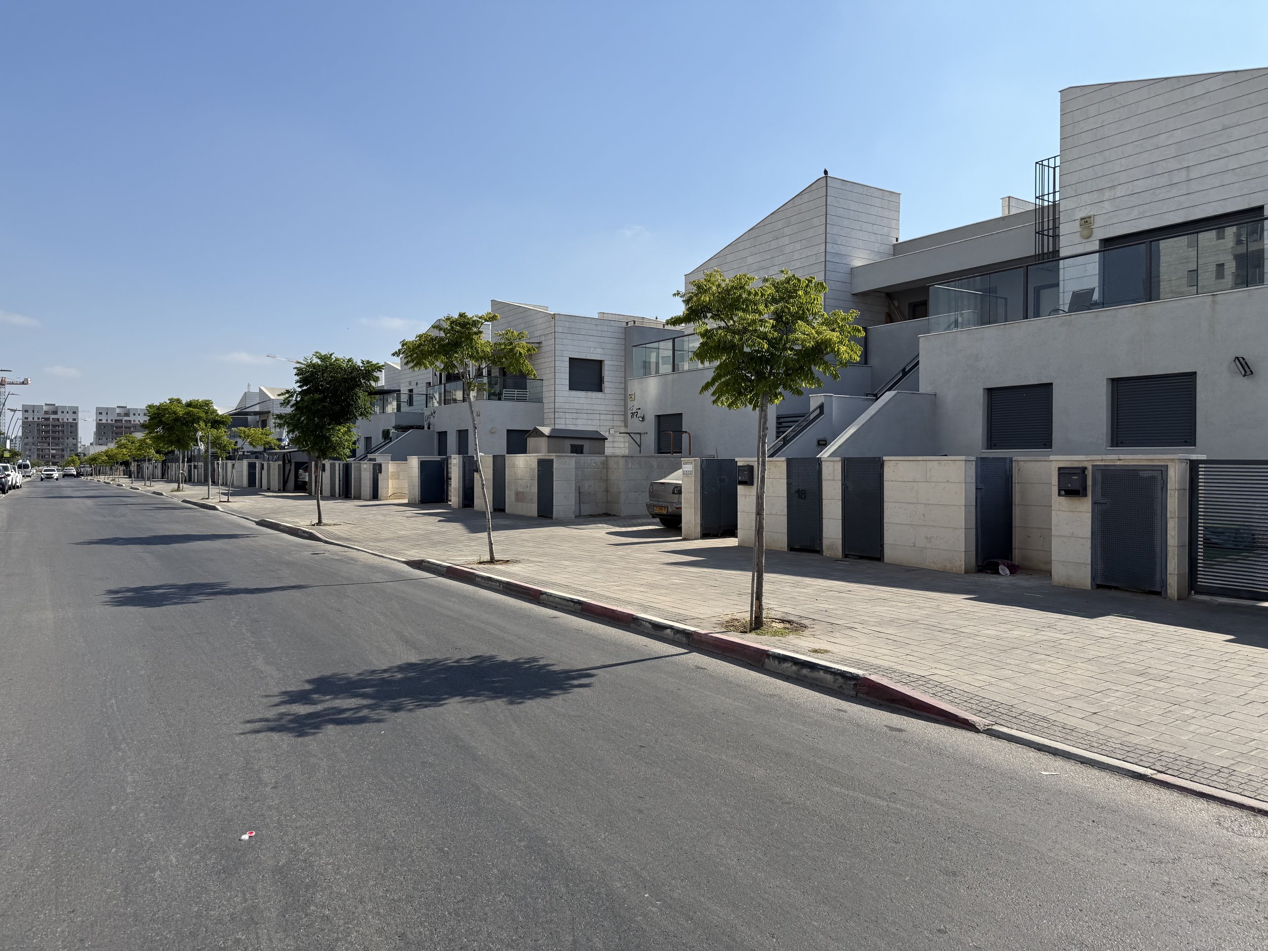 Residential street with modern white houses, trees lining the sidewalk, parked cars, and clear blue sky.