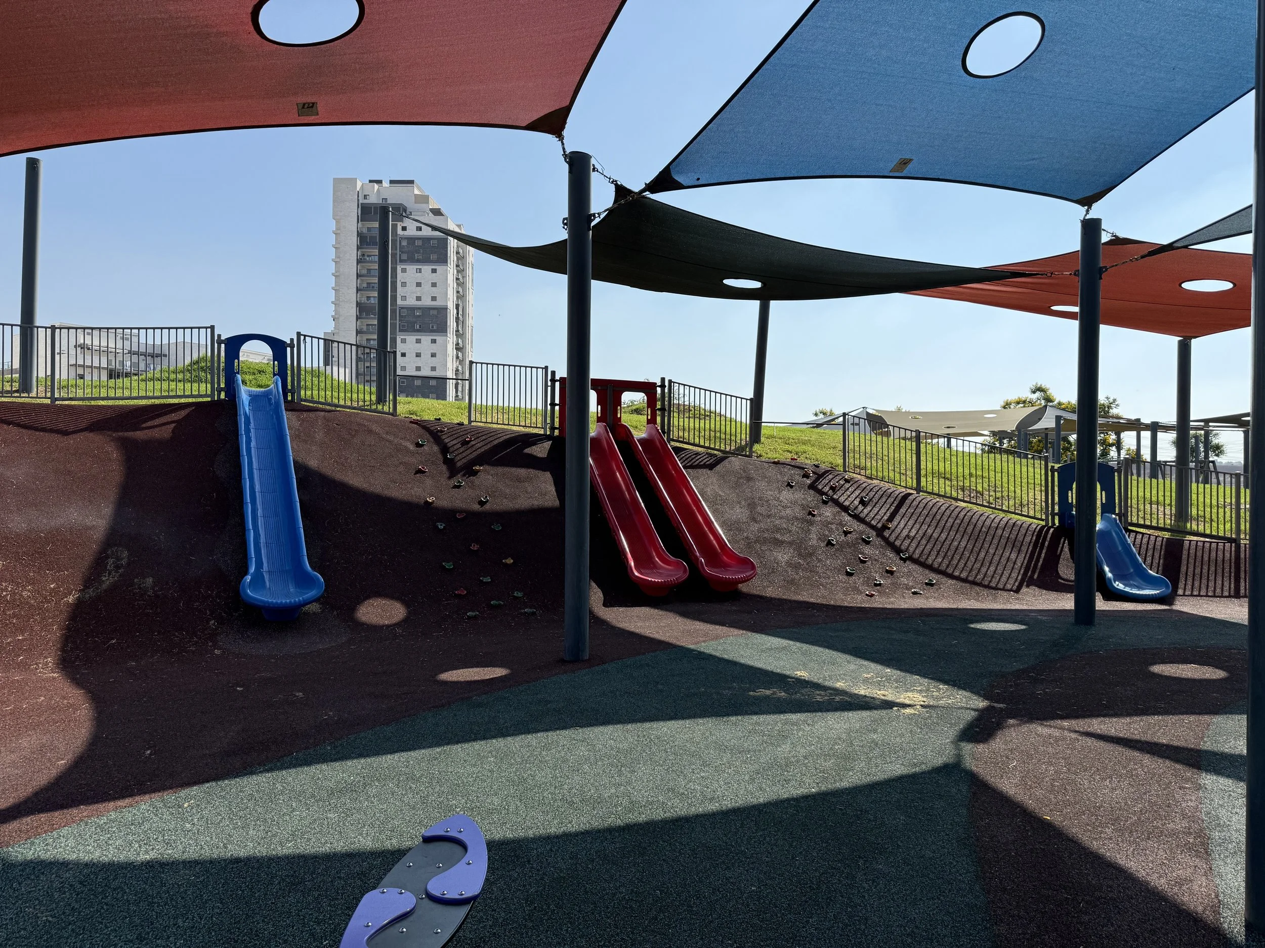 Children's playground with three slides, shaded by colorful fabric canopies, on a sunny day with a city building in the background.