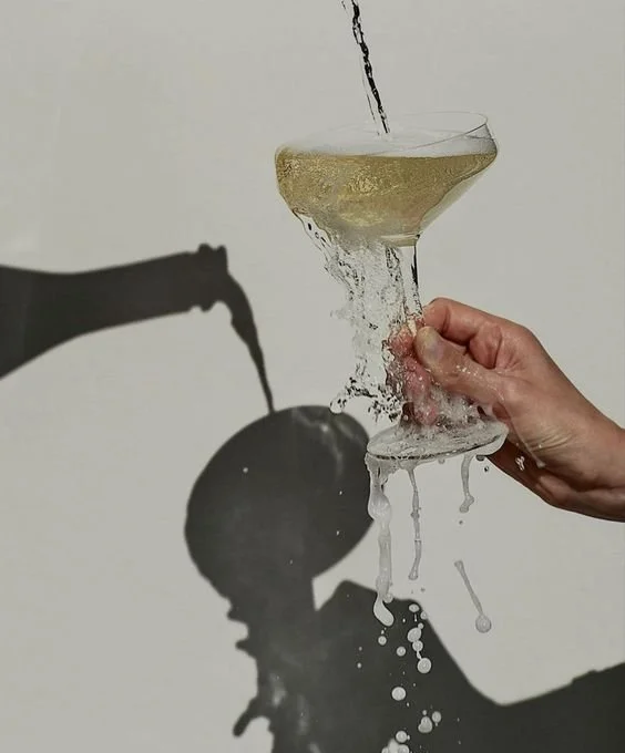 A hand holding a coupe glass with a drink, with water being poured into it, creating a splash and shadow on the wall behind. Burghley Horse Trials After Party.