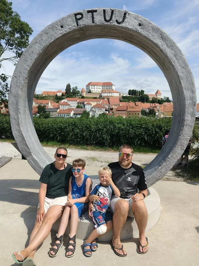 A family of four sitting in front of a large circular sculpture with the word 'PTUJ' at the top. The sculpture frames a view of a historic town with orange rooftops and a castle on a hill in the background. The family is smiling, wearing casual summer clothing and sunglasses.