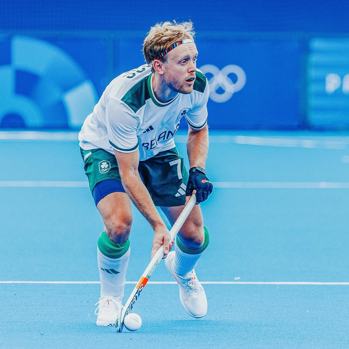A male field hockey player in a white and green uniform, wearing a headband, is bent over with a hockey stick, focusing on the ball on a blue field during a game.