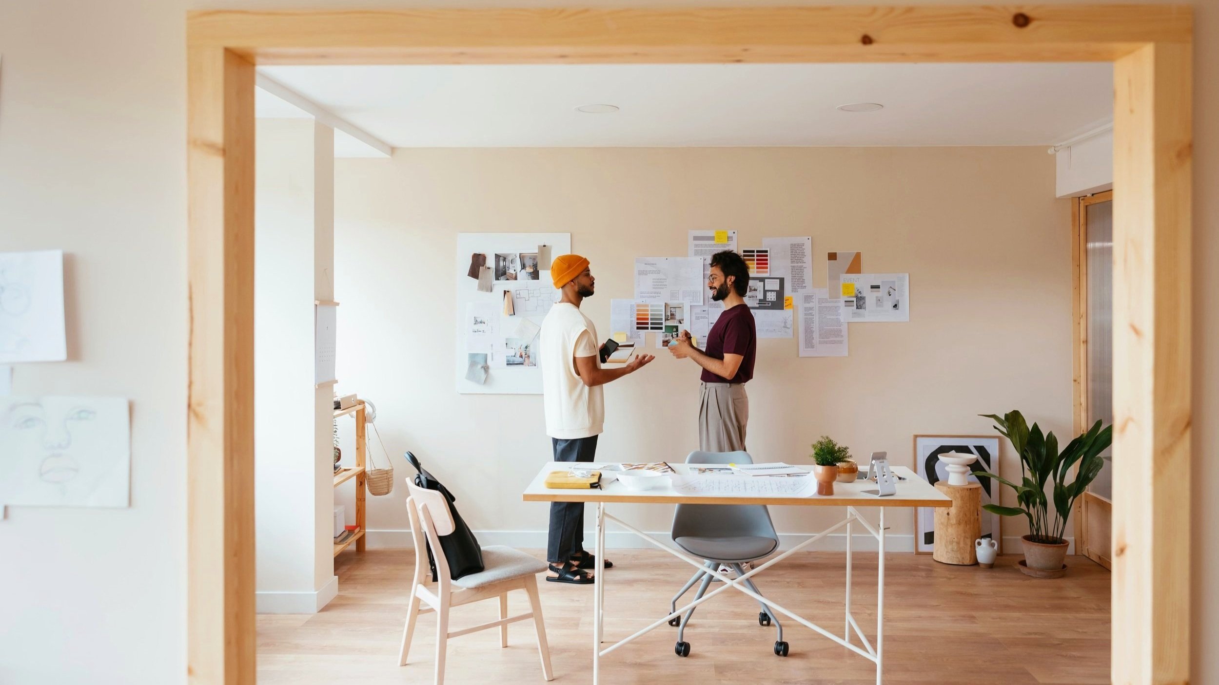 Dois homens conversando em uma sala de escritório com parede de papel de parede com notas e fotos fixadas, mesa com documentos e plantas, decoração de paredes clara e aconchegante.