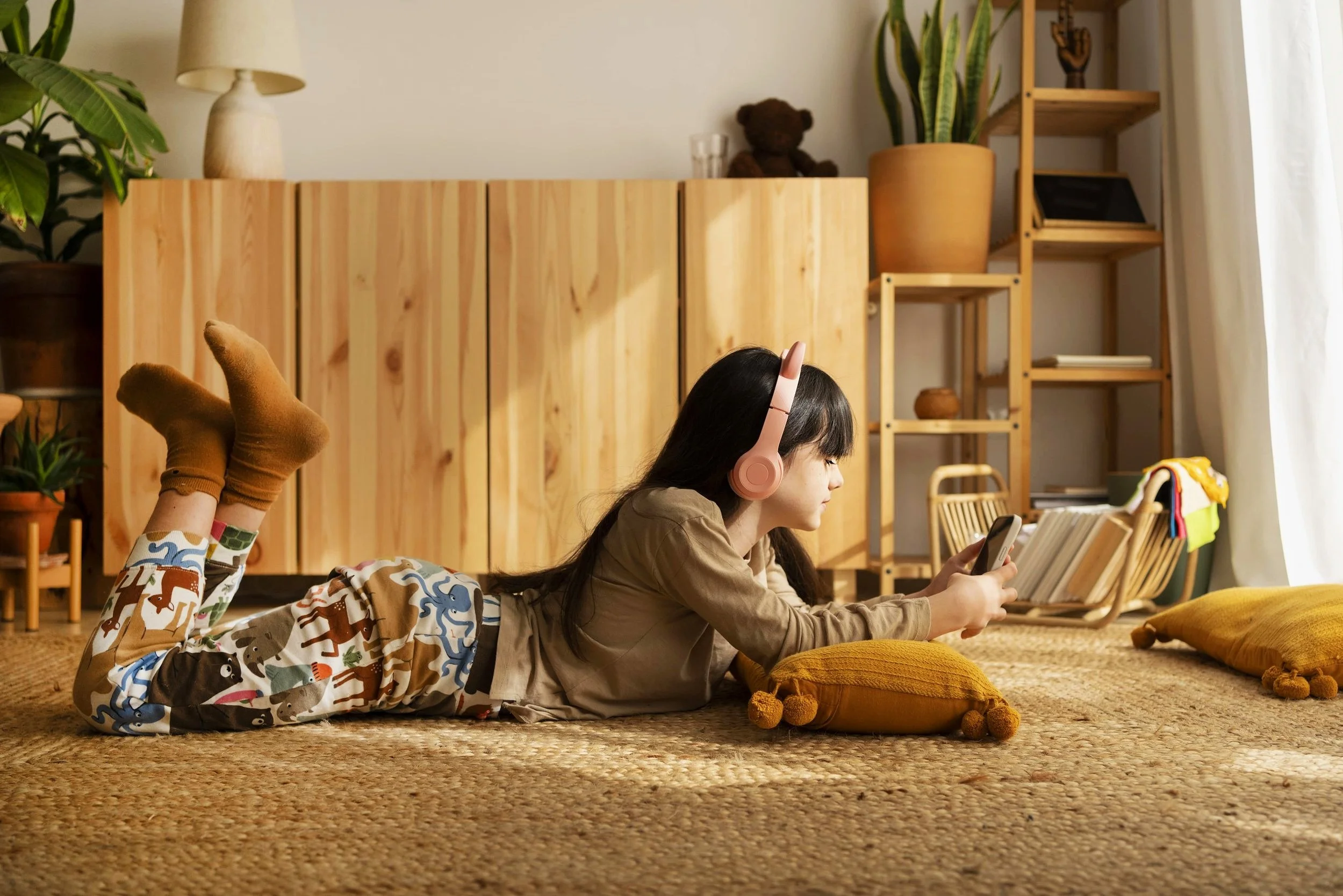 Junges Mädchen im Pyjama und mit rosa Kopfhörern, das im Wohnzimmer auf dem Boden liegt und auf ein Smartphone schaut