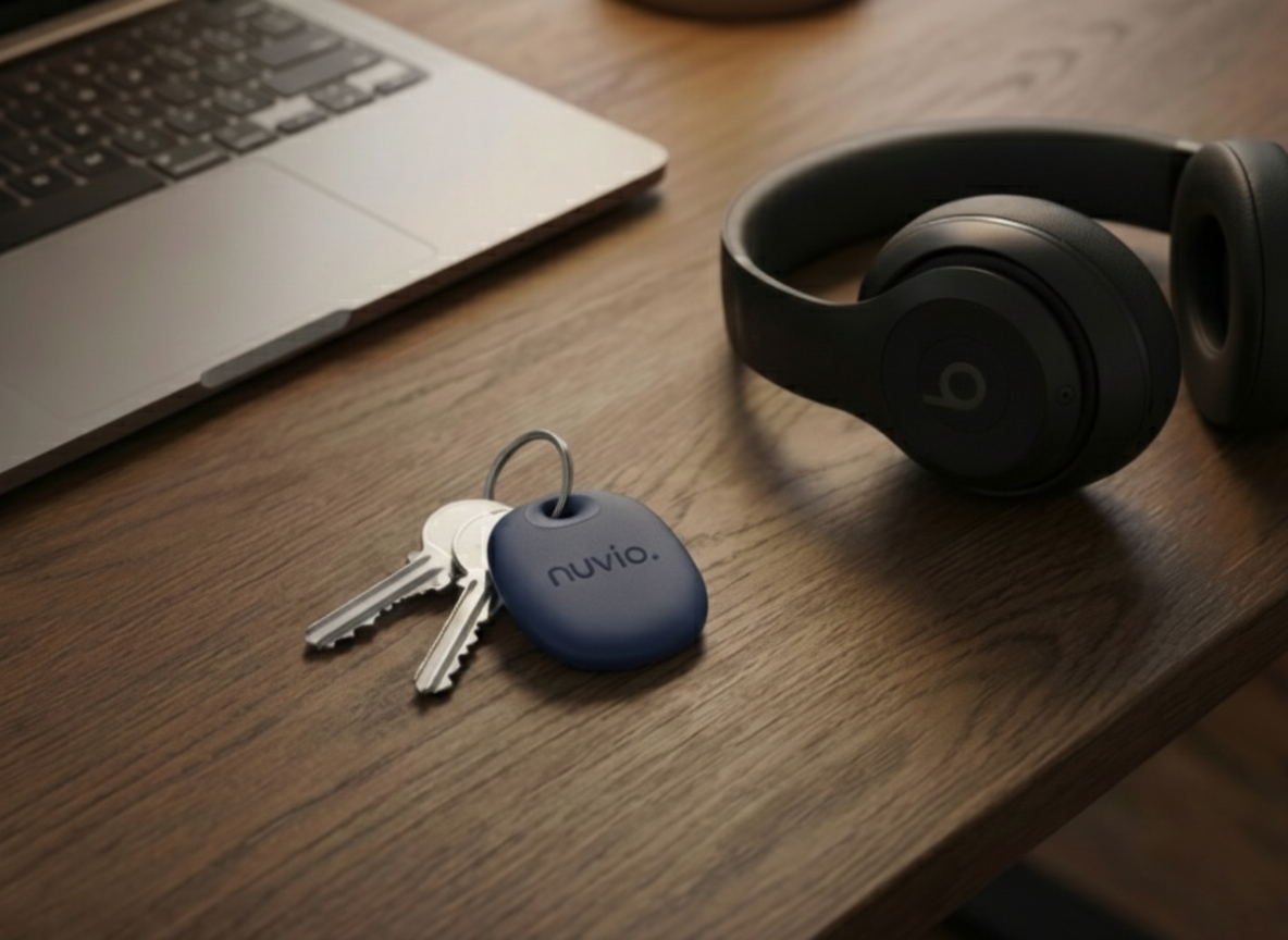 Keys with a blue key cover labeled 'nuvio' traxtag, a pair of black over-ear headphones, and a laptop on a wooden desk.