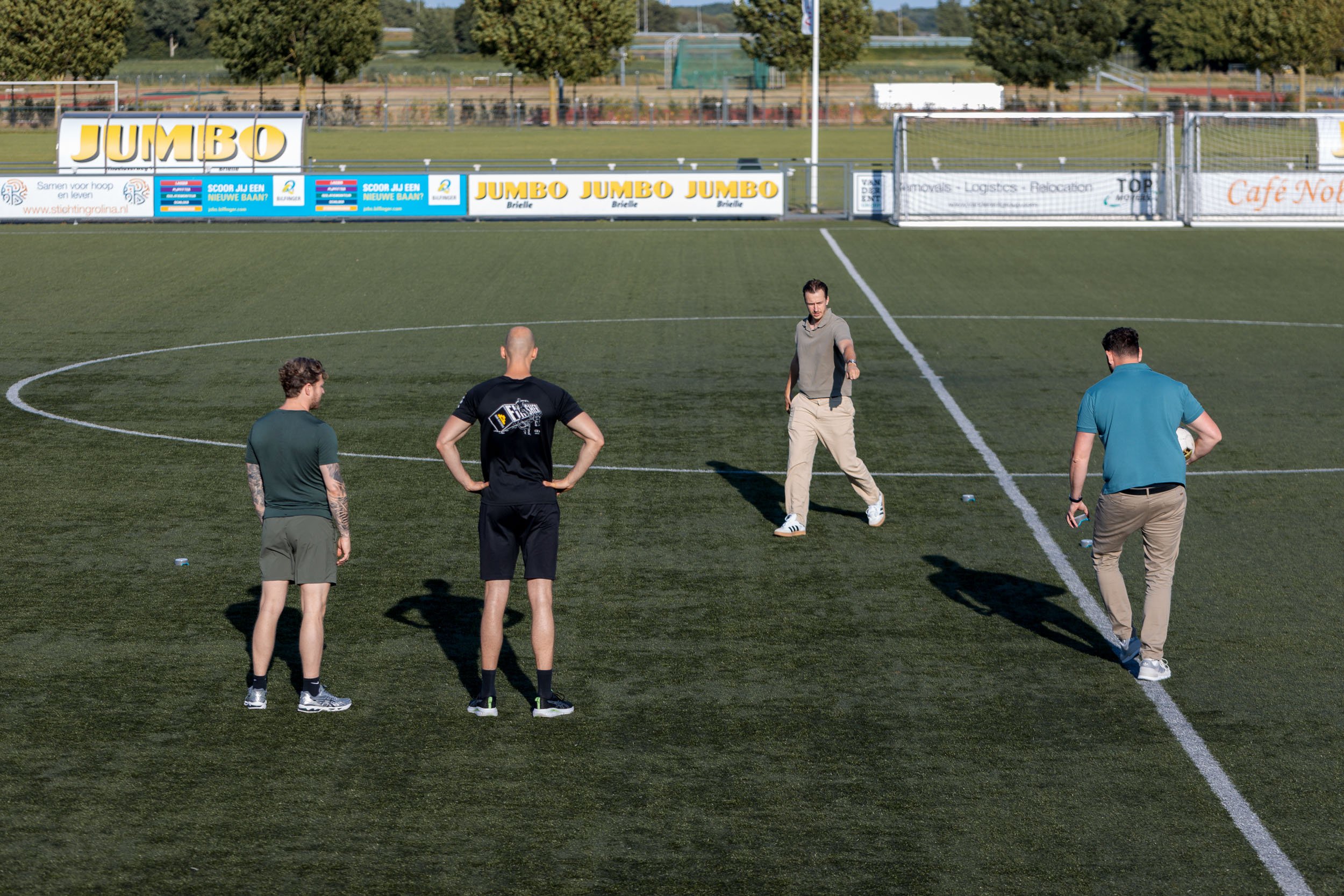 Vier mannen staand op een voetbalveld, waarbij één man iets uitlegt terwijl hij wijst, in helder daglicht. Op de achtergrond zijn reclameborden en bomen zichtbaar.