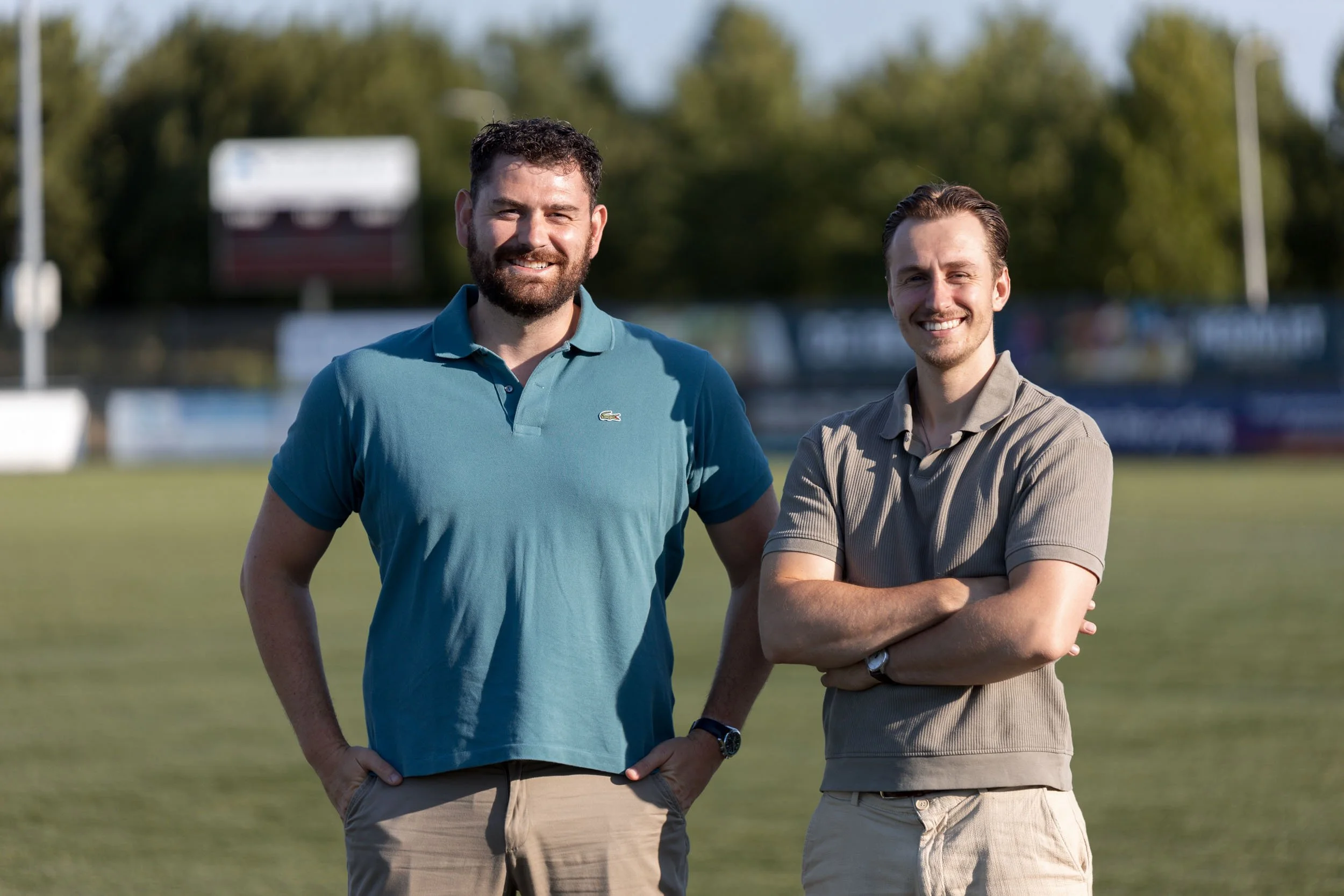Twee lachende mannen op een voetbalveld met bomen en een hek op de achtergrond, één met een blauw poloshirt en de ander met een beige shirt en korte broek.
