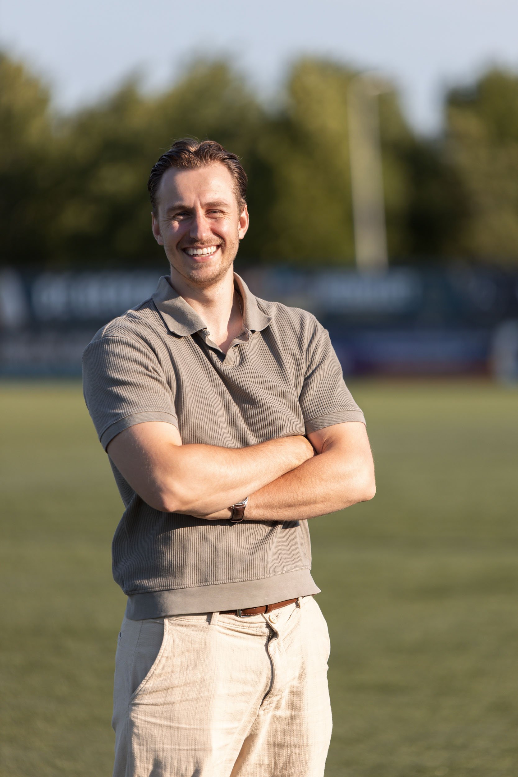 Een lachende man met bruin haar en een baard, staand in de buitenlucht op een grasveld met bomen op de achtergrond. Hij heeft zijn armen gekruist en draagt een beige poloshirt met korte mouwen en lichtgekleurde broek.