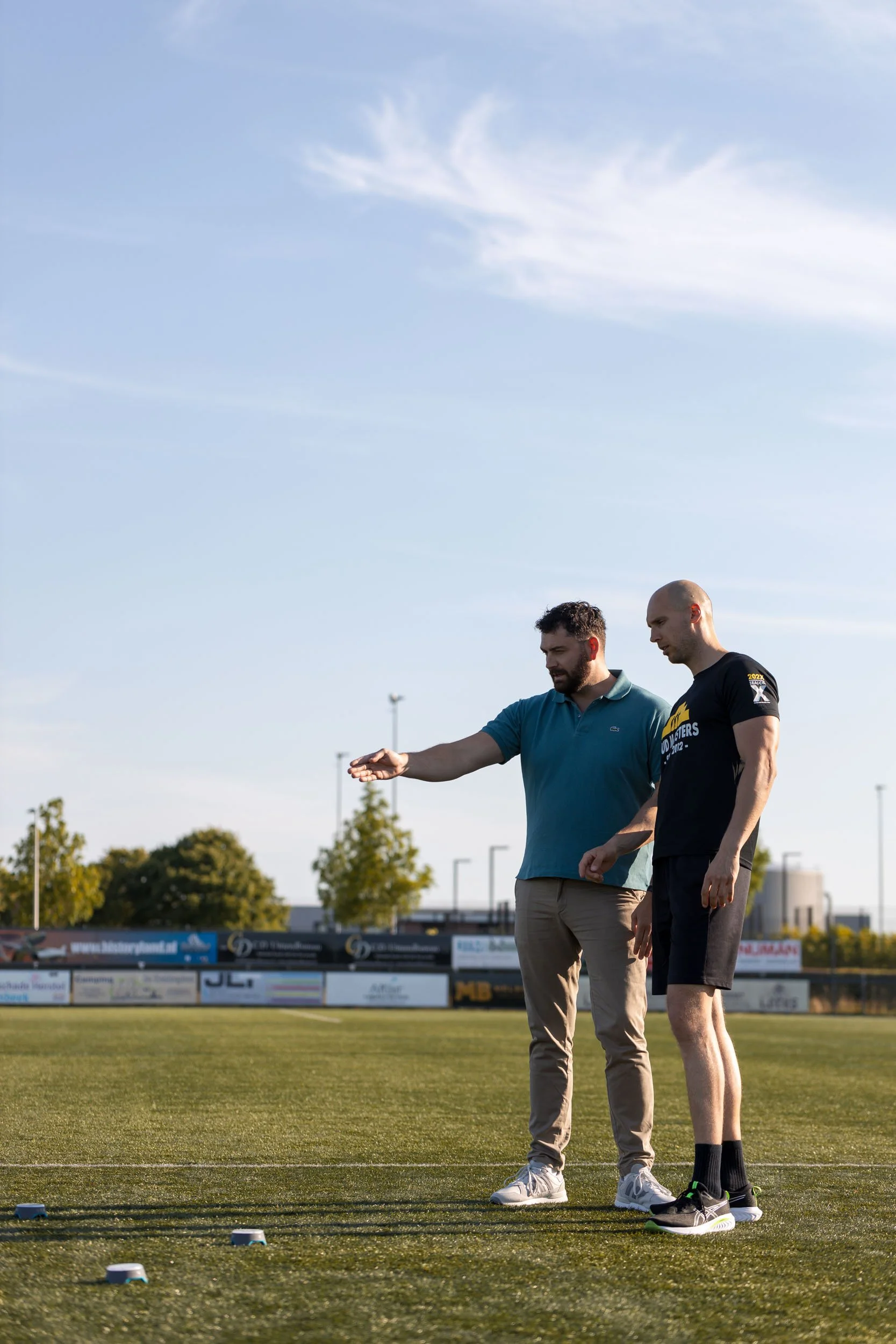 Twee mannen staand op een sportveld, iets besprekend of inspecterend, met een heldere lucht en bomen op de achtergrond.