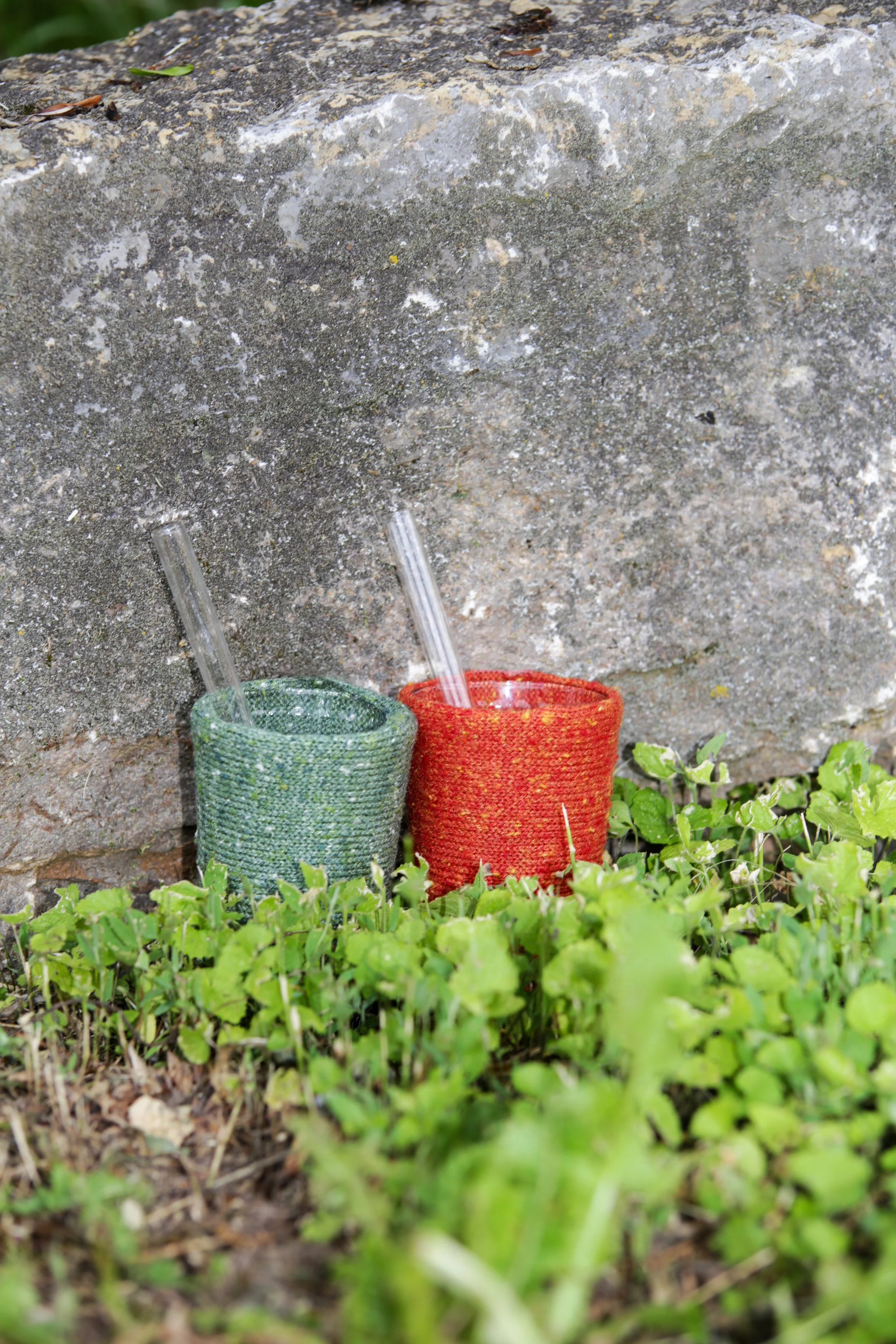 Two colorful knitted cups, one green and one red, with clear straws, placed on the grass near a stone wall.