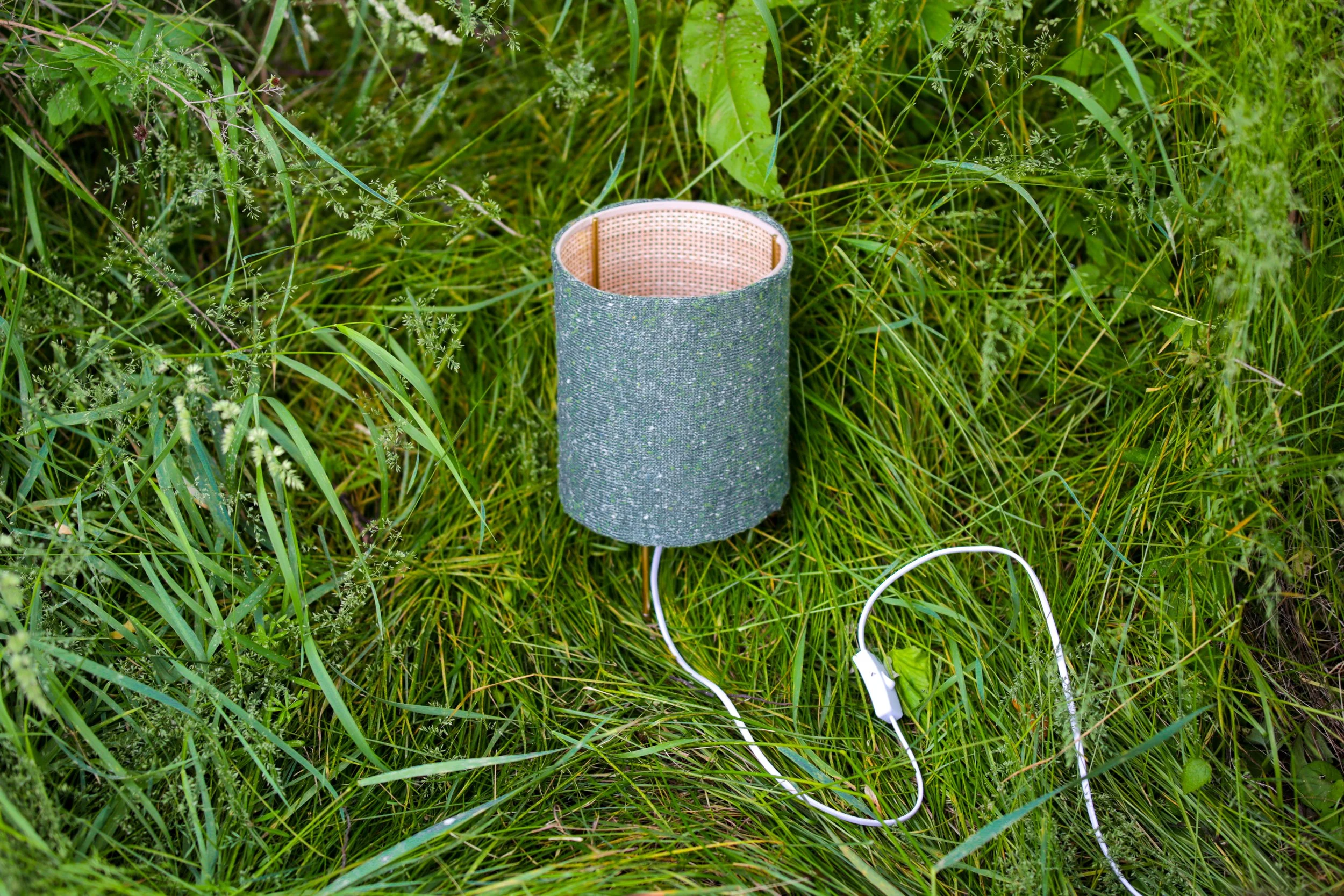 A cylindrical speaker with a textured gray exterior is lying on green grass, with a white charging cable attached nearby.