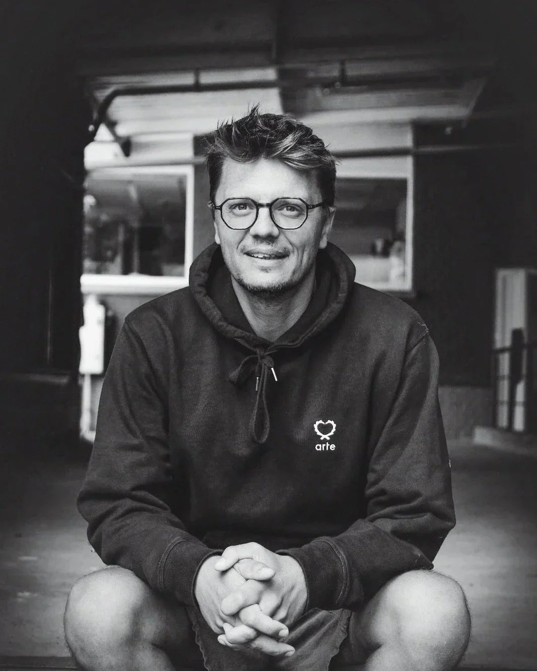 Un homme assis, portant des lunettes et un sweat à capuche noir avec un logo « arte » sur la poitrine, dans un environnement intérieur, photographié en noir et blanc.