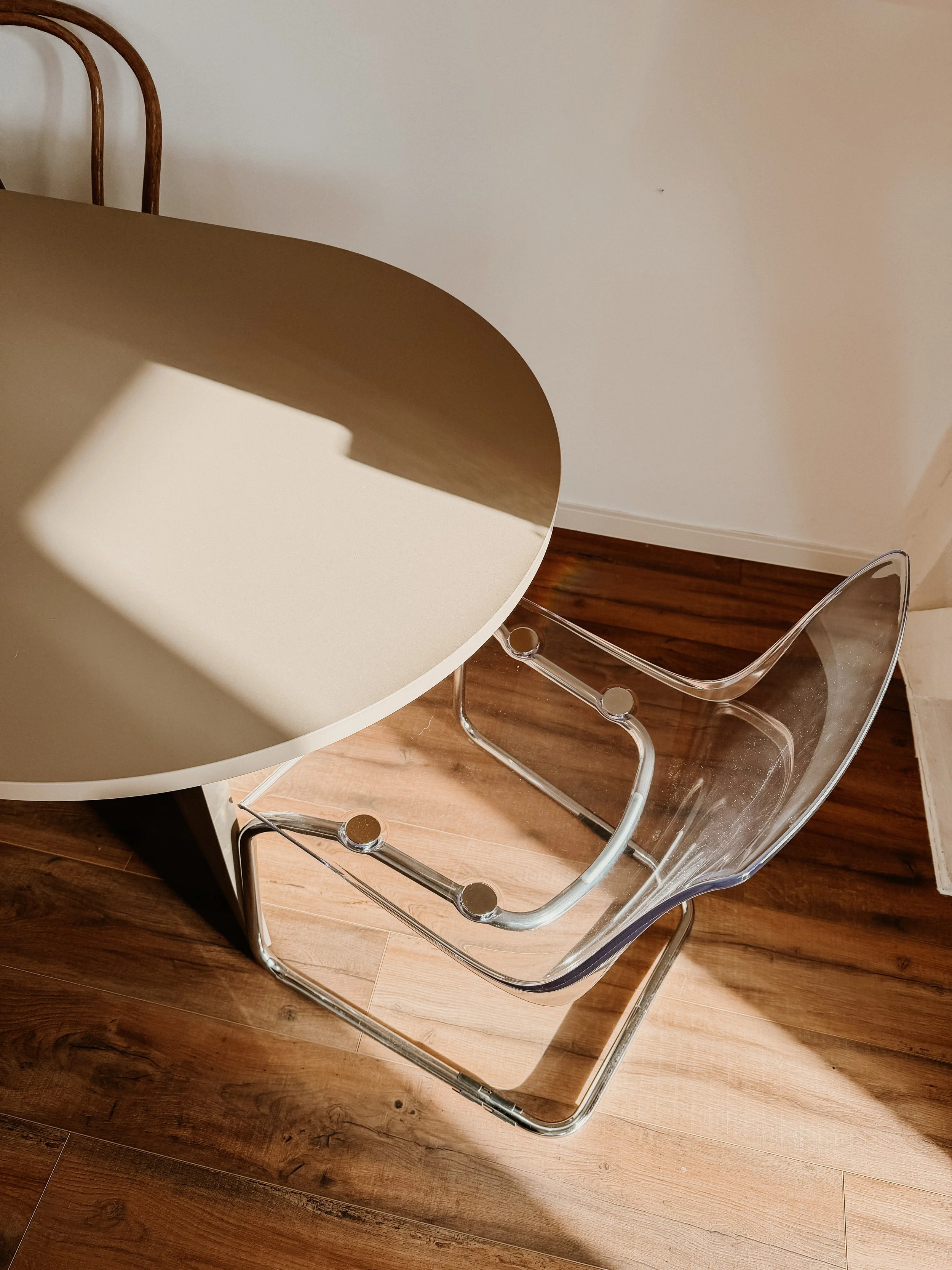Ein transparenter Stuhl mit abgerundeter Sitzfläche und eine runde, beige Tischplatte, alles auf einem Holzboden.