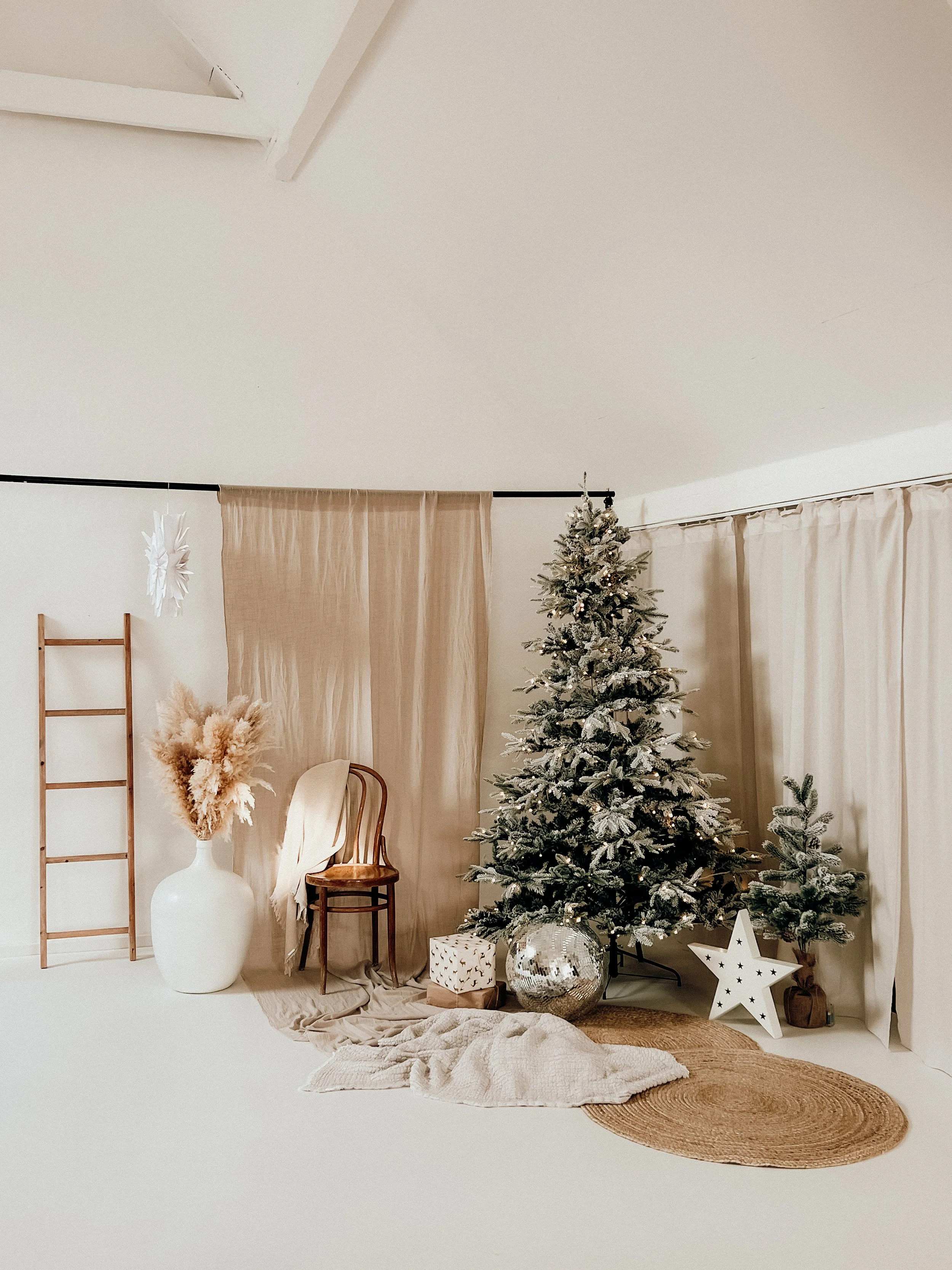 Eine moderne Weihnachtsdekoration mit einem geschmückten Weihnachtsbaum, Geschenken, einer großen weißen Vase mit getrockneten Pflanzen, Holzstühlen, einem kleinen Tisch, Kerzen, einem Stern und einem Läufer auf dem Boden vor einem beige Vorhang.